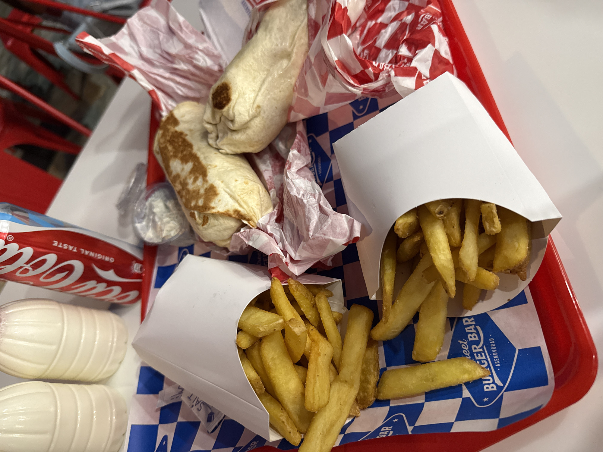 A high-angle shot captures a fast-food meal laid out on a red plastic tray. The tray holds two white paper containers, each filled with golden-brown french fries. Adjacent to the fries are two wrapped items, resembling burritos or kebabs, with lightly toasted tortillas, resting on crumpled red and white checkered paper.
A red Coca-Cola can is partially visible on the left side of the tray, clearly displaying "Coca-Cola" and "ORIGINAL TASTE" in white script. Below the can are two distinct white, ribbed, oval-shaped containers, likely shakers for condiments such as salt or pepper.
The various food items and their packaging are placed on a blue and white checkered paper liner. Visible text on this liner includes "Sweet BURGER BAR" and "ASENOVGRAD", pinpointing the establishment's name and location, consistent with the provided context of Bulgaria. Additional partial text, "SAIT" (possibly 'salt'), and "BAR" are also visible on the paper. The scene suggests a ready-to-eat meal from a casual dining or take-out restaurant, likely indoors due to the bright, consistent lighting.