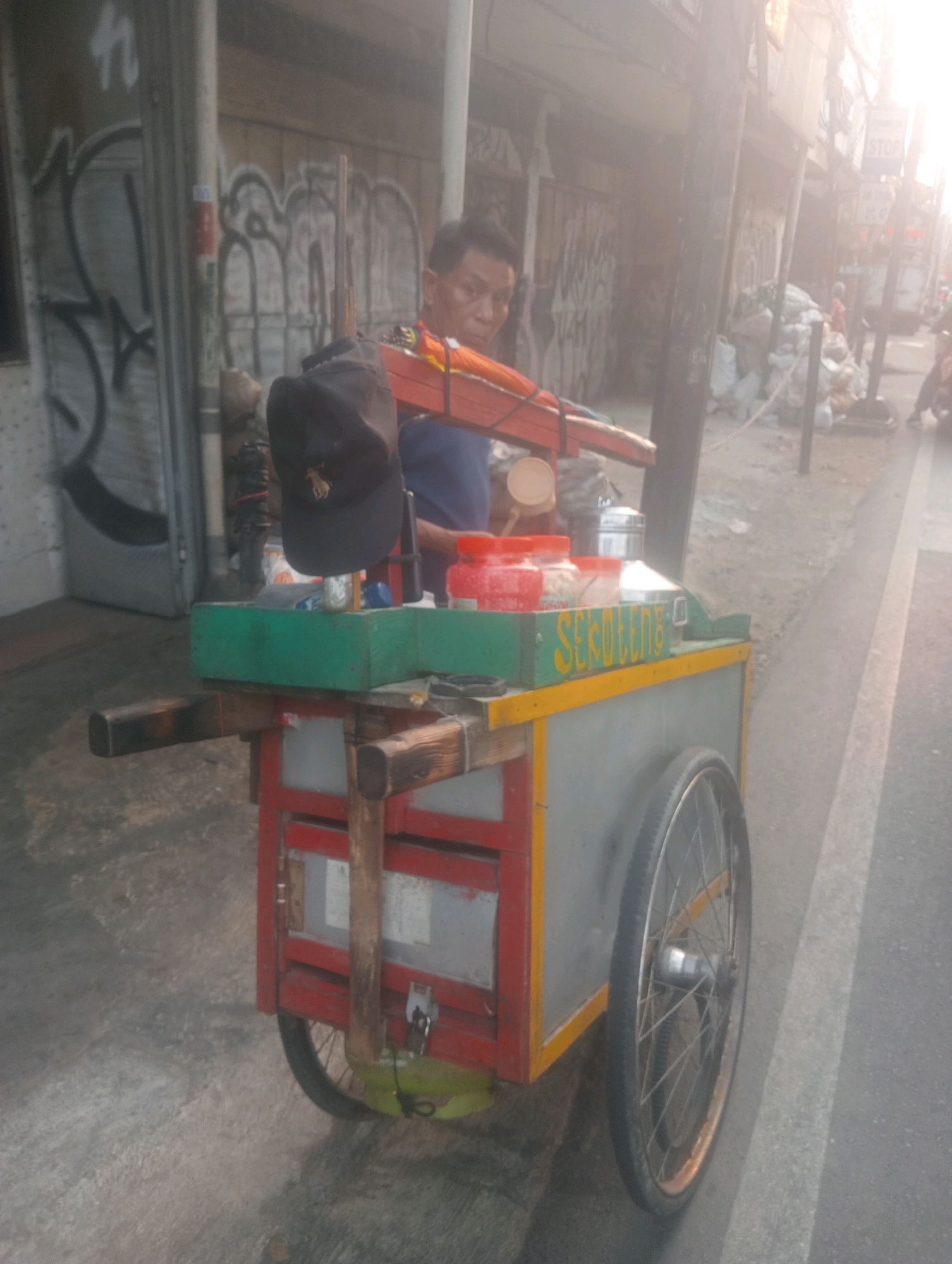 In the city of East Jakarta, Indonesia, a street vendor is seen next to his mobile food cart. The vendor, a middle-aged man wearing a blue shirt, is partially visible, facing to the left of the frame. He has a dark cap perched on his head, positioned on the cart's railing. The cart itself is constructed with a gray body and a red frame, featuring a green top shelf with yellow trim. On this shelf, several containers are arranged: two red plastic containers, a silver metal container, and a clear plastic container. The cart also displays the word "SEDULEN" in yellow lettering on the green section. A large black tire is attached to the cart's left side, and another is partially visible on the right. The background shows a street scene with graffiti on a wall, a paved sidewalk, and a road with white lane markings. In the distance, trash bags are piled up, and other figures are blurred, suggesting a bustling urban environment. The lighting indicates it is daytime, with some sun glare creating a hazy effect.