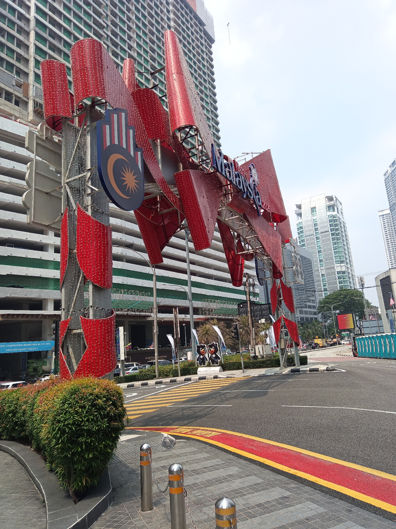 An exterior, eye-level, medium shot captures a large, red, sculptural landmark with the word "Malaysia" emblazoned across it, set against a backdrop of city buildings and a bright sky. The landmark is composed of multiple red, curved elements, some cylindrical and others more angular, adorned with small, white, reflective lights or studs. A Malaysian flag, featuring a crescent moon and star on a blue background, is prominently displayed on the structure.
In front of the landmark, a street with yellow and white road markings, including a pedestrian crossing, is visible. A line of manicured green bushes borders the paved walkway in the foreground. The walkway itself is made of grey cobblestones and is punctuated by silver bollards with yellow reflective bands, connected by chains.
Behind the landmark, a partially constructed tall building with a grid-like facade dominates the mid-ground. Further back, several modern skyscrapers with glass exteriors rise against the sky. Other elements in the scene include traffic lights, street lamps, parked cars, and a porta-potty unit on the right side of the road. The overall environment suggests an urban setting in Kuala Lumpur, Malaysia, likely in a commercial or public area given the prominent signage and infrastructure. There are no people clearly visible in the frame.