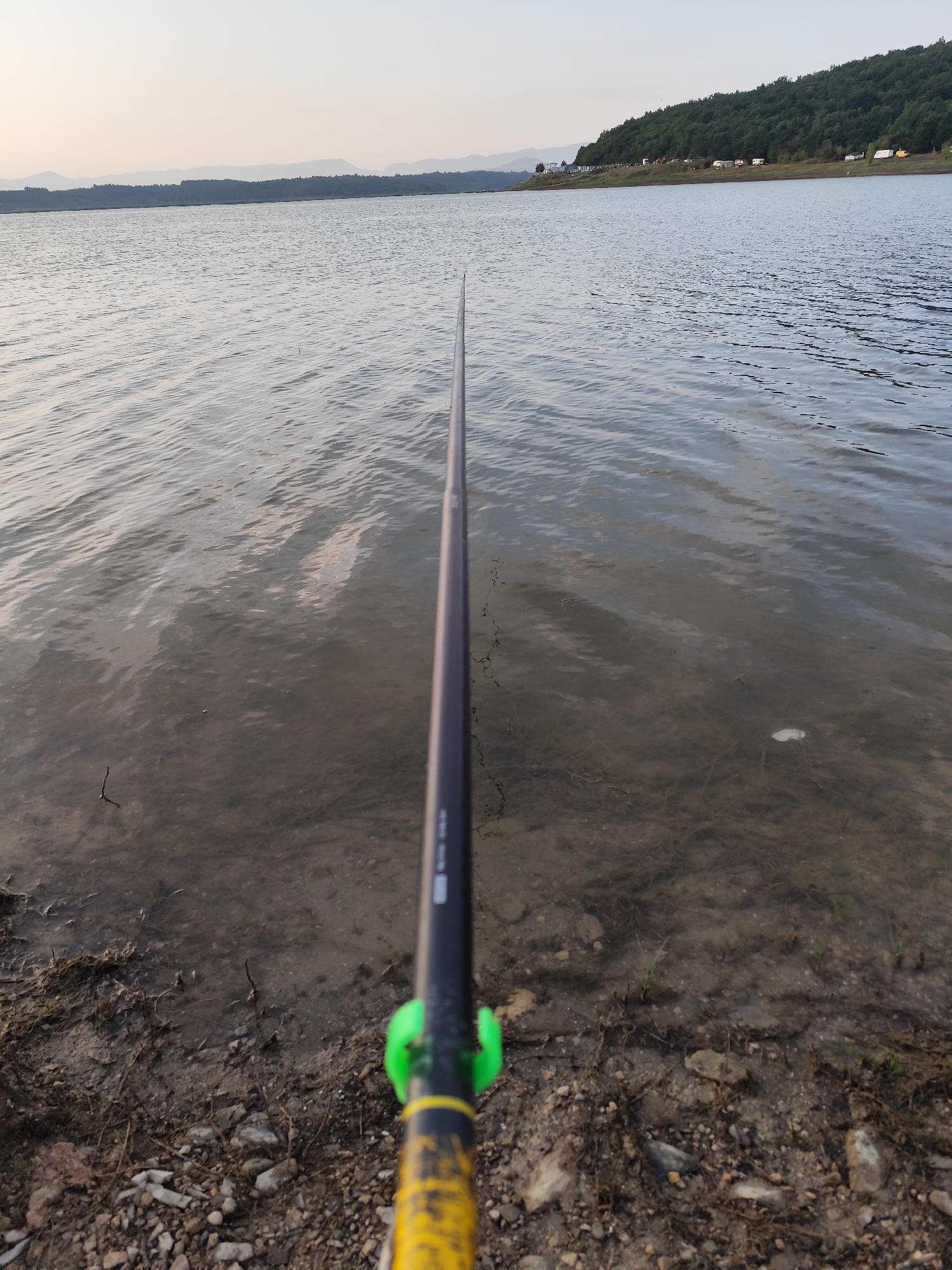 The image depicts a serene natural scene with a body of water in the foreground and background. A fishing rod extends from the bottom right corner of the frame, angling towards the water's surface. The water is calm, with gentle ripples reflecting the soft, hazy light of what appears to be early morning or late evening. The shoreline is composed of pebbles and dirt, with some sparse vegetation. In the distance, a treed hillside rises, dotted with what appear to be small structures, possibly campers or recreational vehicles, suggesting a lakeside recreational area. The sky is a pale, diffused hue, with distant, muted mountain ranges visible on the horizon. The overall impression is one of peaceful solitude and outdoor recreation. The location is identified as the Ogosta Reservoir, near the city of Montana, Bulgaria.