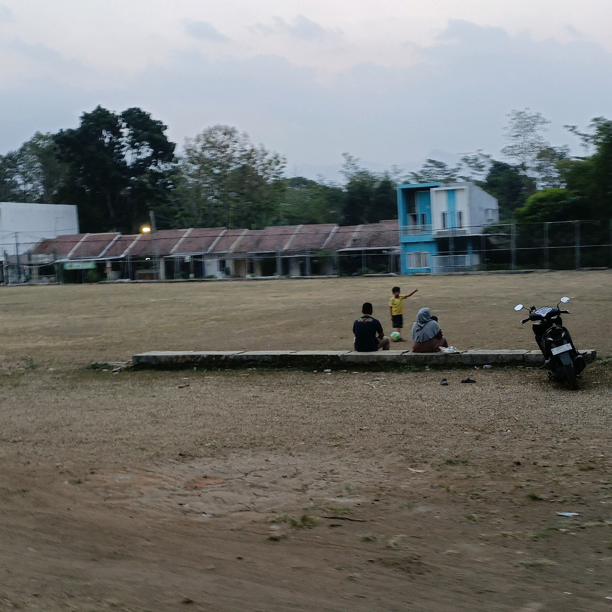The image depicts an outdoor scene at what appears to be a sports field or open area, likely in Salatiga, Indonesia, given the provided context. The ground is predominantly dry, light brown earth, with some patches of sparse green vegetation. In the background, a long, low building with a tiled, sloping roof, possibly housing or administrative facilities, runs horizontally across the frame. Adjacent to this structure is a more modern, multi-story building with a distinctive blue and white color scheme. A chain-link fence delineates the perimeter of the field.
In the mid-ground, several individuals are present. A young boy wearing a yellow t-shirt stands in the field, his right arm raised, seemingly gesturing or playing. A soccer ball is visible near him. Two adults are seated on a low concrete wall or ledge running across the field. One is wearing a dark blue shirt, and the other, a woman, is wearing a grey hijab and a darker colored garment. A black motorcycle is parked on the right side of the frame, facing away from the viewer. The sky above is overcast with a pale, muted light, suggesting it is either early morning or late evening. The overall atmosphere is quiet and relaxed.