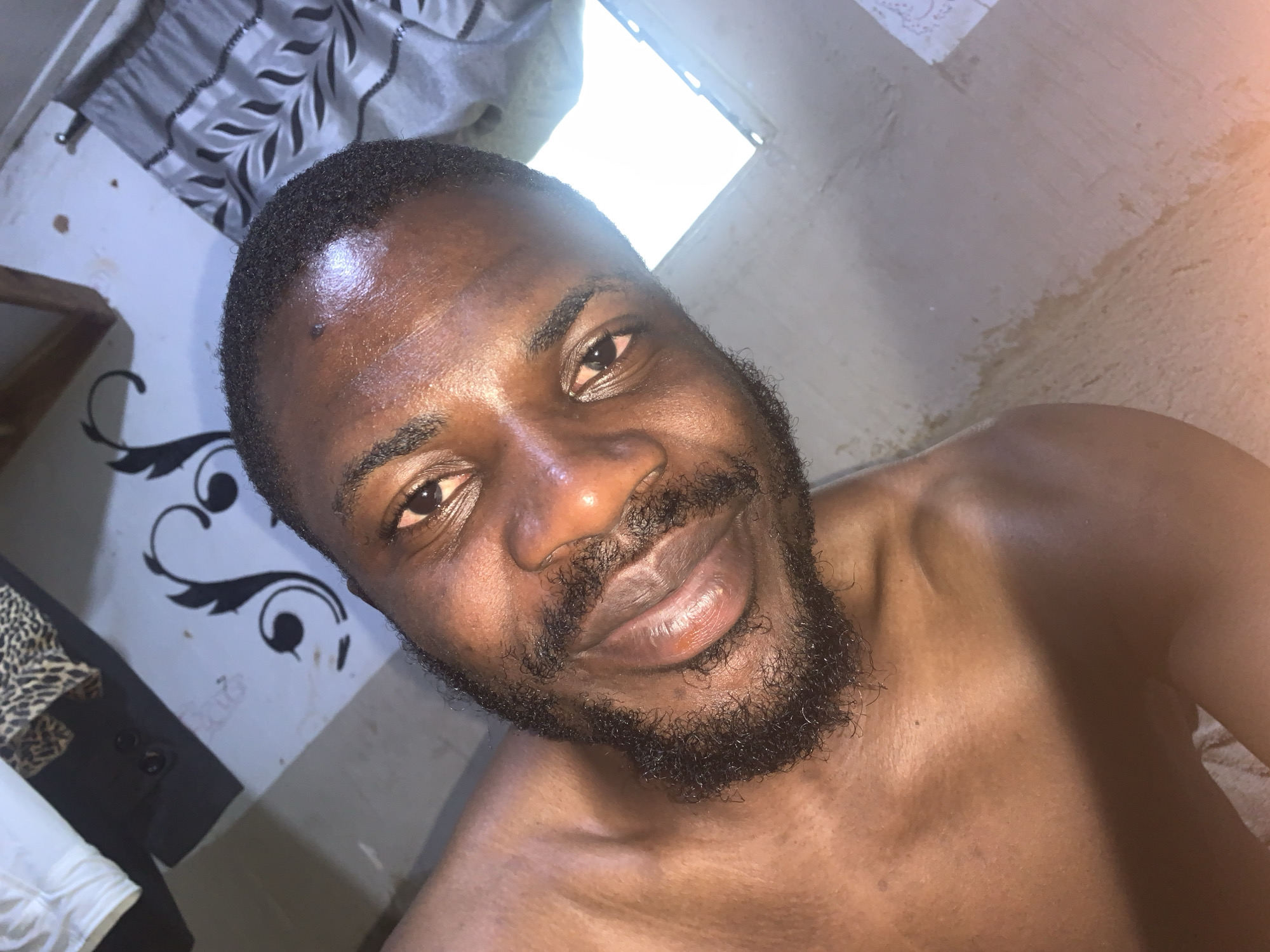 A close-up, overhead shot features a Black male, appearing to be in his late 20s or early 30s, with a short beard and dark hair. He is shirtless and looking directly at the camera with a slight smile. His skin is dark and appears slightly oily, reflecting the light. The background shows a tiled wall with a decorative black stencil pattern on the left, and a light-colored curtain with a striped pattern on the right. The lighting suggests an indoor environment, possibly a room in Rafin Par, Nigeria, with light coming from an overhead source or a window. The overall impression is a casual, personal photograph.