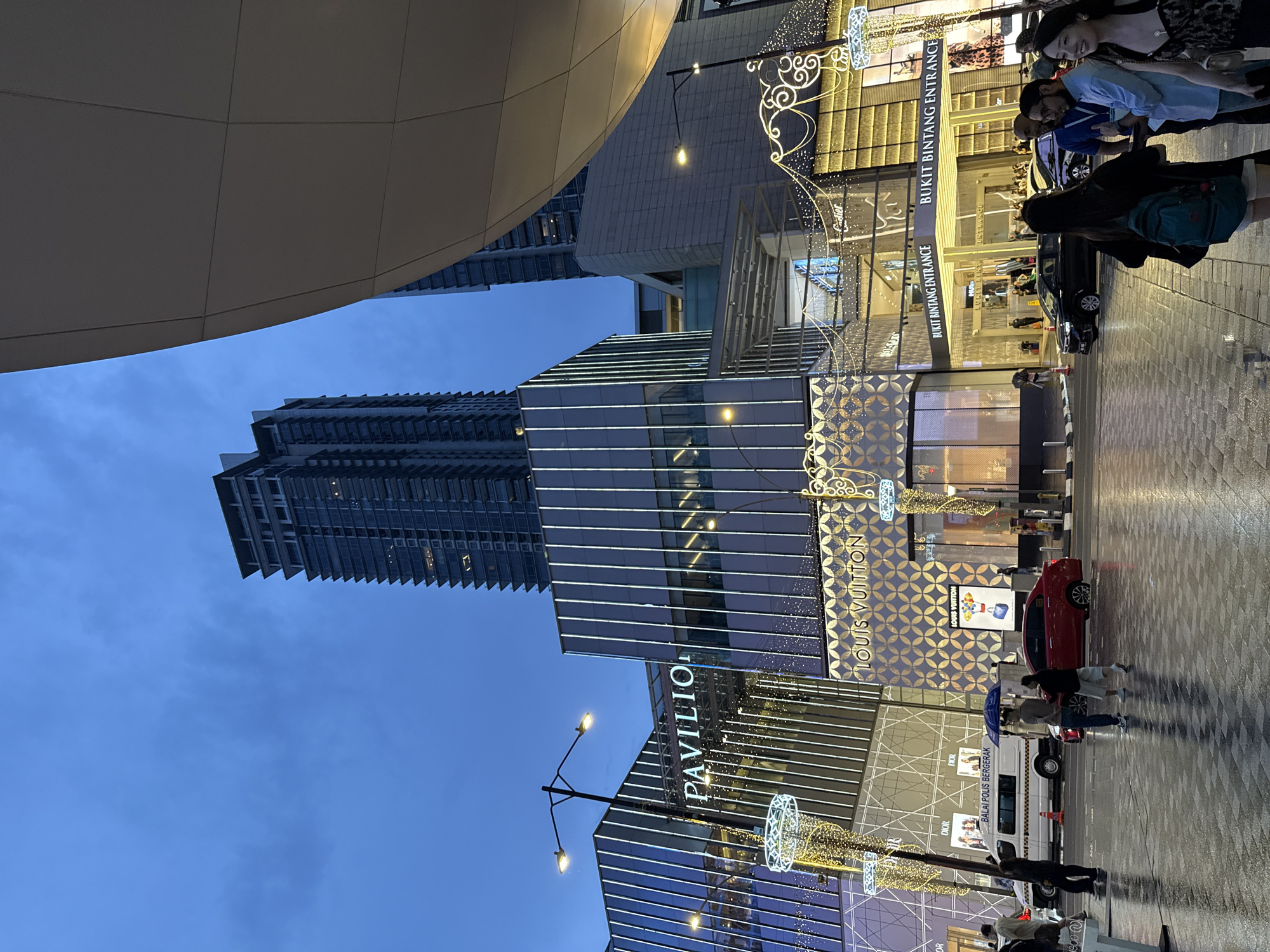 The image shows an evening view of the Pavilion Kuala Lumpur shopping mall in Kuala Lumpur, Malaysia. The architecture is modern, with a towering skyscraper in the background. Street lights and decorative lights adorn the area. People are seen walking along the pavement near the mall's entrance, which has signage for Bukit Bintang Entrance. The fronts of luxury stores such as Louis Vuitton, Dior and Cartier are visible. A police patrol vehicle is also present on the street.