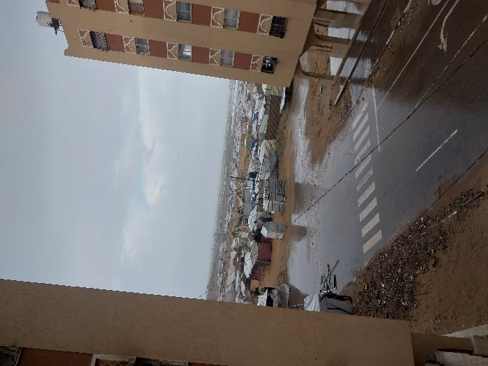 The image presents an expansive, elevated view of a temporary settlement in Khan Yunis, Palestinian Territory, under an overcast sky, with the ground visibly wet from recent rain. In the immediate foreground, a wet asphalt road stretches horizontally, featuring white-striped crosswalk markings. The road is bordered by saturated, muddy ground and debris.
To the left, a partial view of a multi-story, light brown building is visible, while a similar but more complete multi-story building frames the right side of the image. This right building features decorative patterns above its windows and an elevated ground floor supported by pillars, with a white water tank visible on its roof.
Beyond the road and muddy terrain, a vast, densely populated area of temporary shelters extends into the distance. These shelters are constructed from various materials, including white and dark tarpaulins, plastic sheeting, and fabric, indicating an extensive encampment. Numerous puddles and standing water are visible amidst these structures. A single individual, dressed in dark clothing, stands next to a partially collapsed or dismantled tent structure on the left side of the road. A black utility cable traverses the scene horizontally above the road. The sky is uniformly gray and overcast, contributing to the muted lighting of the scene.