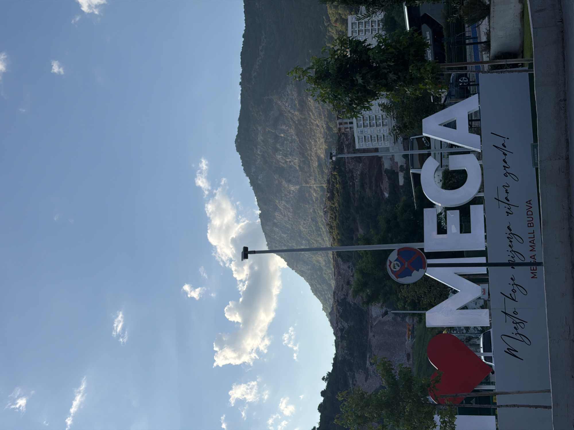 The image displays a large "I LOVE MEGA" sign in Budva, Montenegro, with a red heart and white letters. The sign is situated in front of a mountainous backdrop and a modern building complex. The sky is blue with scattered clouds. Below the "I LOVE MEGA" sign, there is an inscription in what appears to be Montenegrin script, followed by "MEGA MALL BUDVA". A traffic sign with a red circle and a diagonal line through a person's head is visible to the right of the sign. Trees line the foreground, and a street or road is present in the lower part of the image.