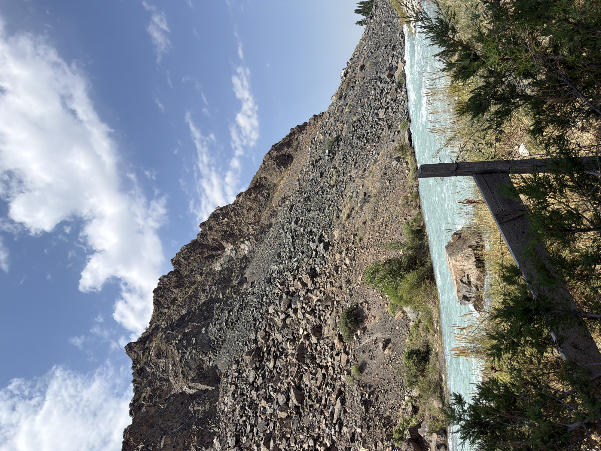 An eye-level, wide shot captures a bright, sunny day in a natural mountainous environment. A fast-flowing, turquoise river dominates the lower half of the frame, its waters churning as it moves from left to right. On the right bank of the river, a weathered wooden fence post stands diagonally, partially obscured by the dense green branches of a conifer in the foreground. The riverbank is lined with dry grasses and a few small green bushes.
The left side of the image is dominated by a steep, rocky mountainside that rises sharply from the river. The slope is covered in loose scree and large boulders, with sparse patches of dry vegetation clinging to the rock face. The peak of the mountain is sharp and jagged. The sky above is a clear, vibrant blue, punctuated by scattered, fluffy white clouds. The overall impression is one of a rugged and untamed natural landscape.