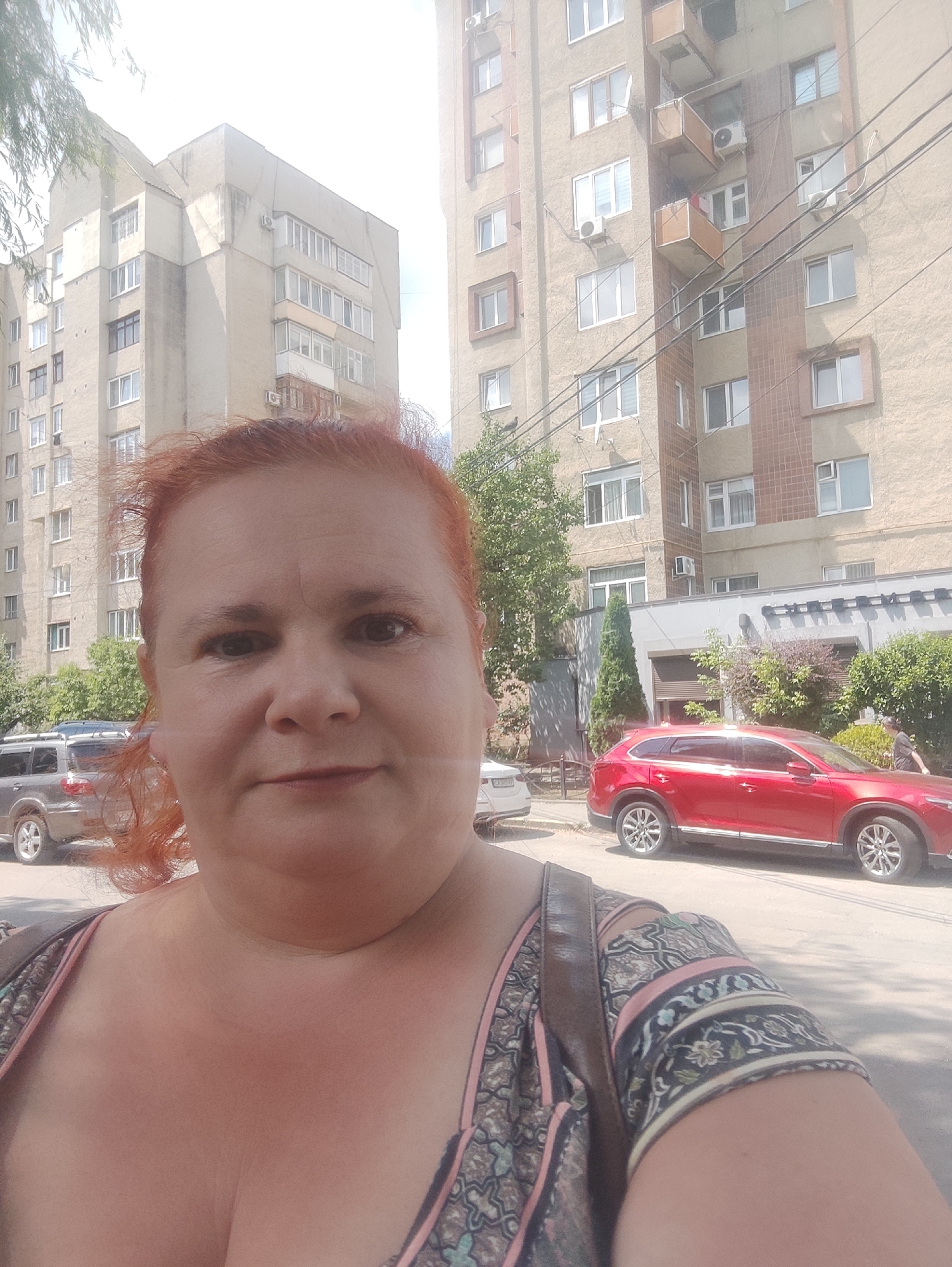 A woman with red hair is taking a selfie outdoors. She is in the foreground, and behind her, two apartment buildings are visible, along with parked cars and trees. The apartment buildings appear to be in a residential area. One building is taller and has many windows, balconies, and air conditioning units. The other is shorter and has a simpler facade. A red SUV is parked on the street in front of the buildings. The overall impression is of a sunny day in an urban environment. There is no specific indication in the image to suggest it is in Chernivtsi, Ukraine.