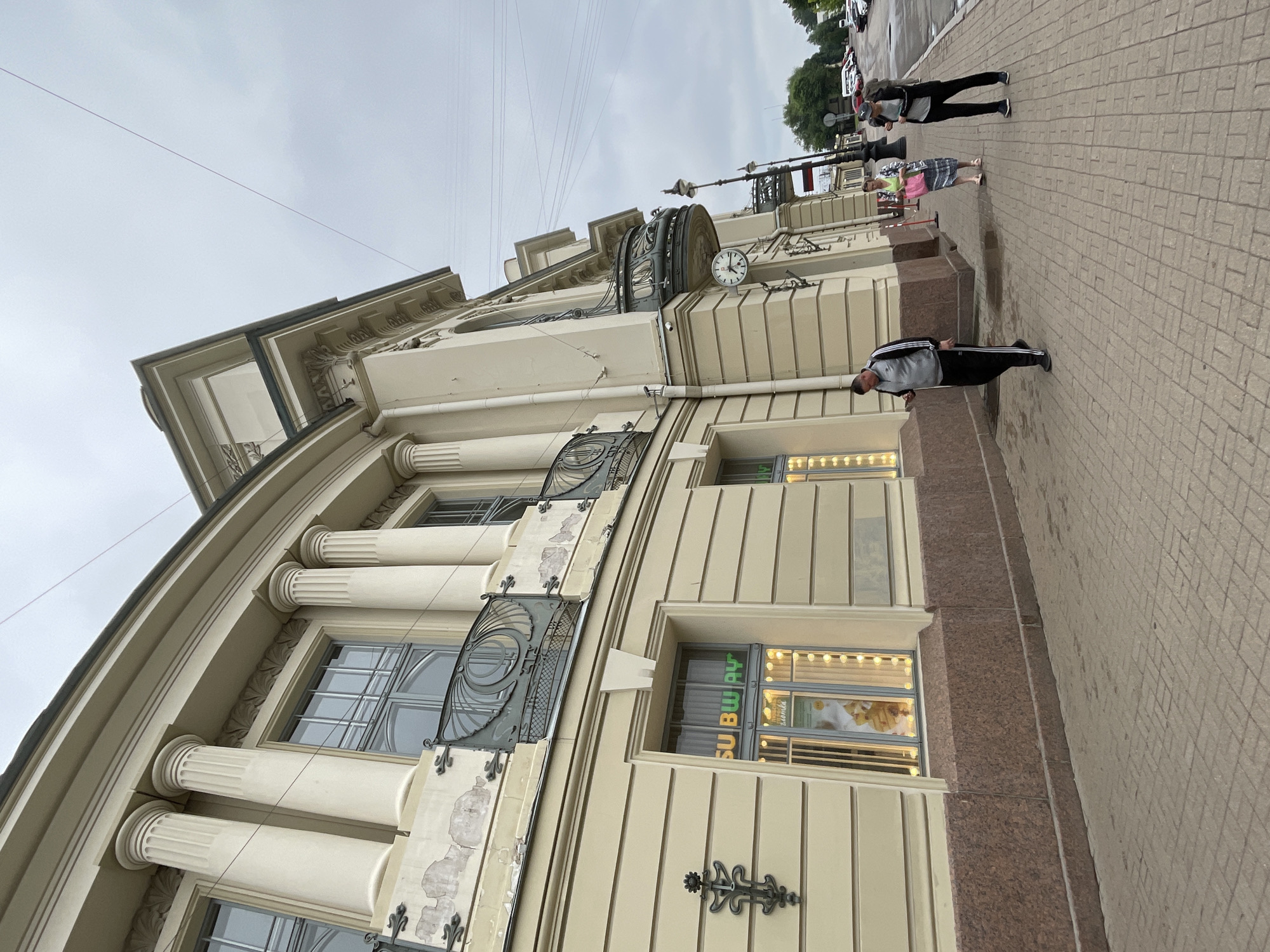 The image shows a building with classical architectural elements, including columns and ornate balconies, on a cloudy day in Saint Petersburg, Russia. A Subway restaurant is visible on the ground floor. Three people are walking on the sidewalk in front of the building. The man closest to the camera is wearing a grey tracksuit and walking away from the viewer, looking at his phone. A woman in a floral skirt and lime green top is walking towards the viewer, carrying a pink bag. Another woman, wearing a dark jacket and cap, is walking away from the viewer, carrying a backpack. The street is paved with brickwork, and cars are parked on the side of the road in the background.