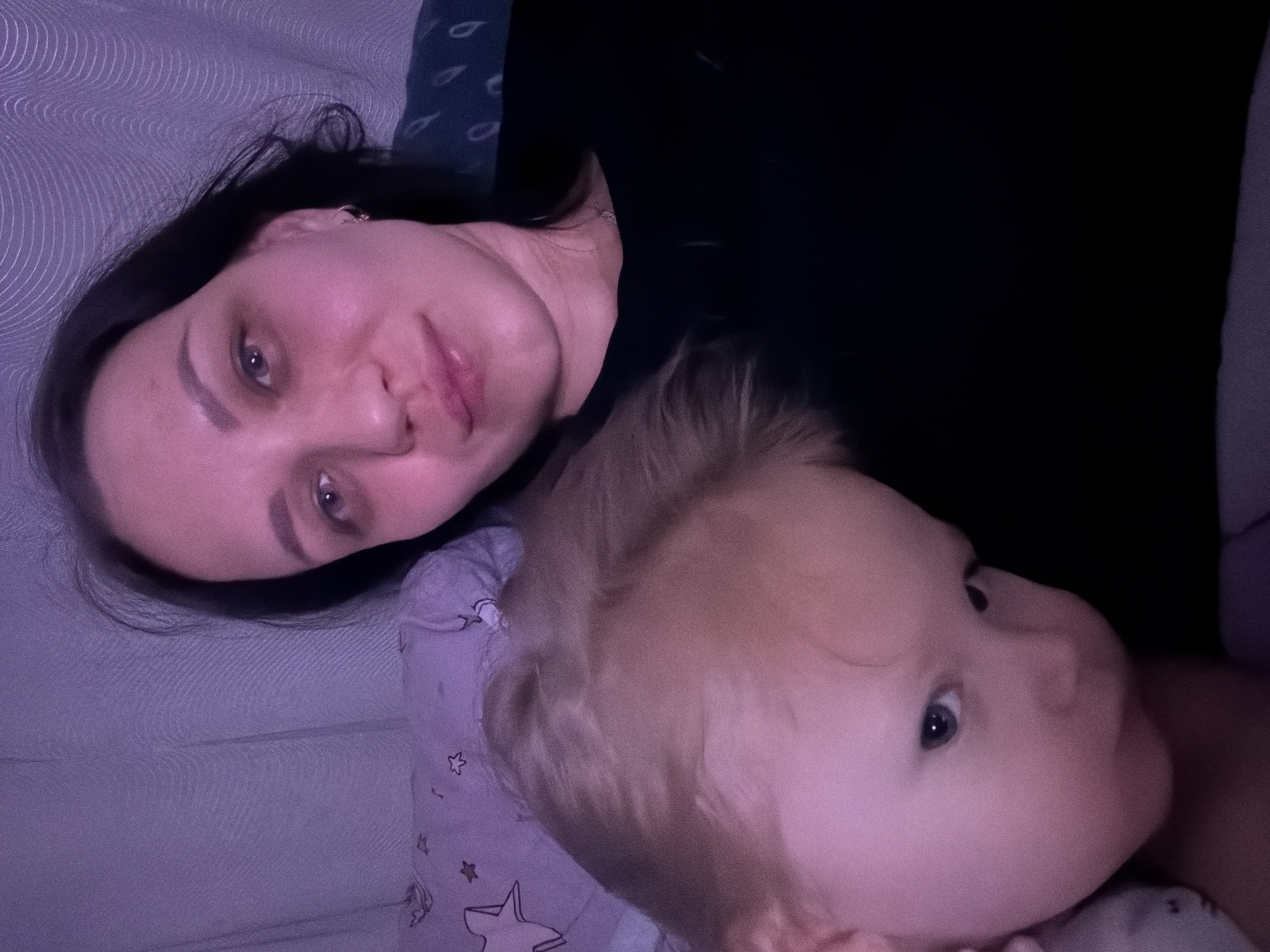 A woman and a young child are in a dimly lit room, possibly a bedroom. The woman is looking towards the camera, and the child is looking away. The background appears to be a textured wall or curtains. The lighting is low, with a purple tint, suggesting artificial light. There are no visible indicators of the location within the city of Perm, Russia.