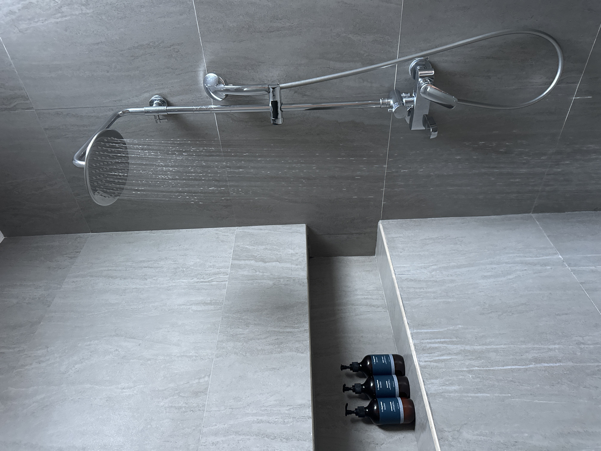 The image shows the interior of a shower room. The walls and floor are covered in large, gray tiles with a textured surface. A chrome shower fixture is mounted on the wall, consisting of a large, round showerhead and a curved overhead pipe connected to a water valve with a flexible hose for a hand shower. Water is flowing from the showerhead. In a recessed niche on the floor are three bottles with pump dispensers. The bottles are dark in color and have a blue label. The location is potentially in Sukawati, Indonesia, based on the request. No people are visible in the image.