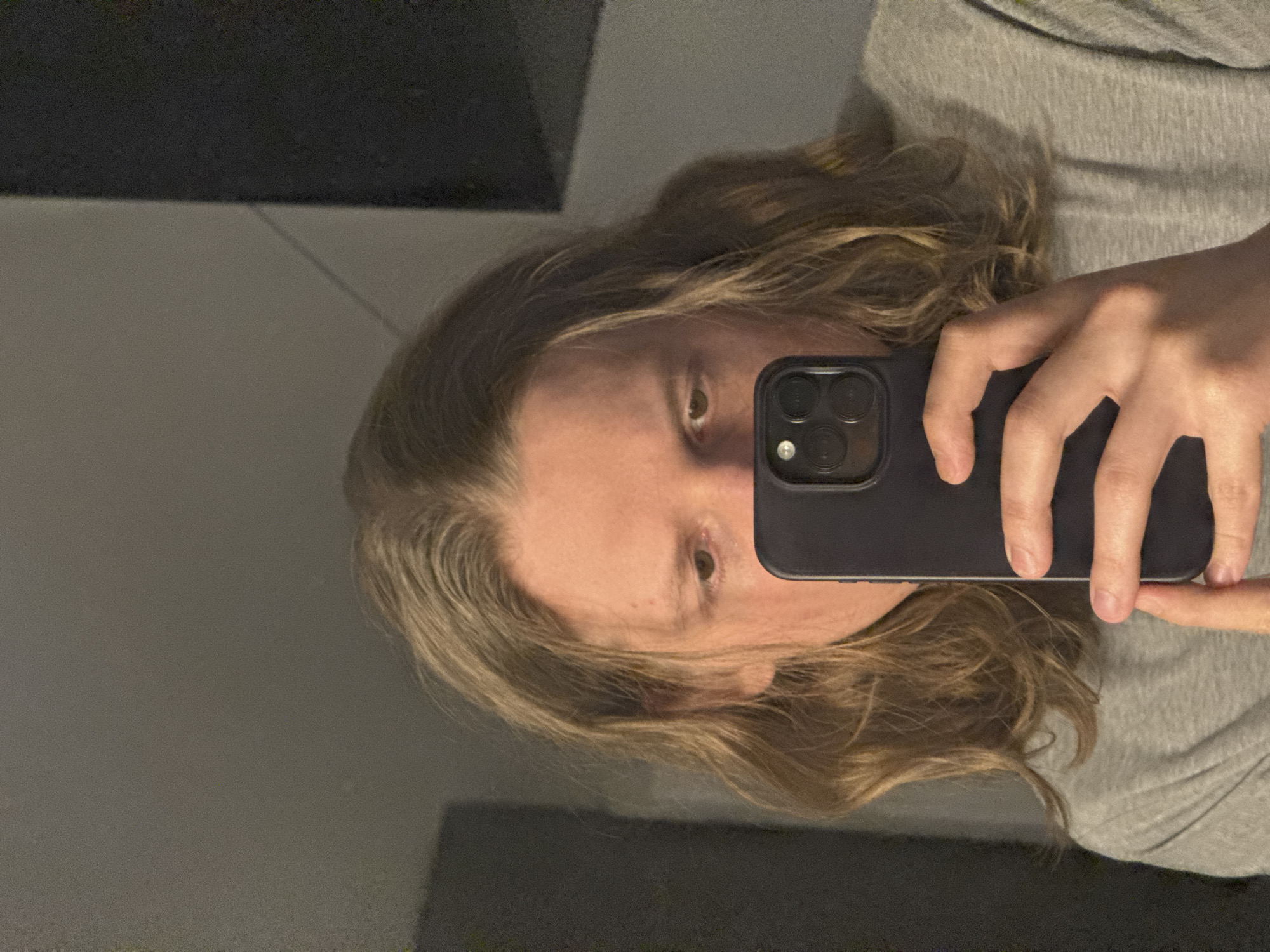 A person with medium-length, wavy, light brown hair is taking a selfie in what appears to be an indoor setting. They are holding a dark-colored smartphone in their right hand, with their thumb and index finger visible. The person is wearing a grey t-shirt. The background is dimly lit and appears to be a wall with some indistinct shapes, possibly furniture or structures, and a dark vertical element on the right side. The lighting is somewhat grainy. The location is Unknown, Unknown.