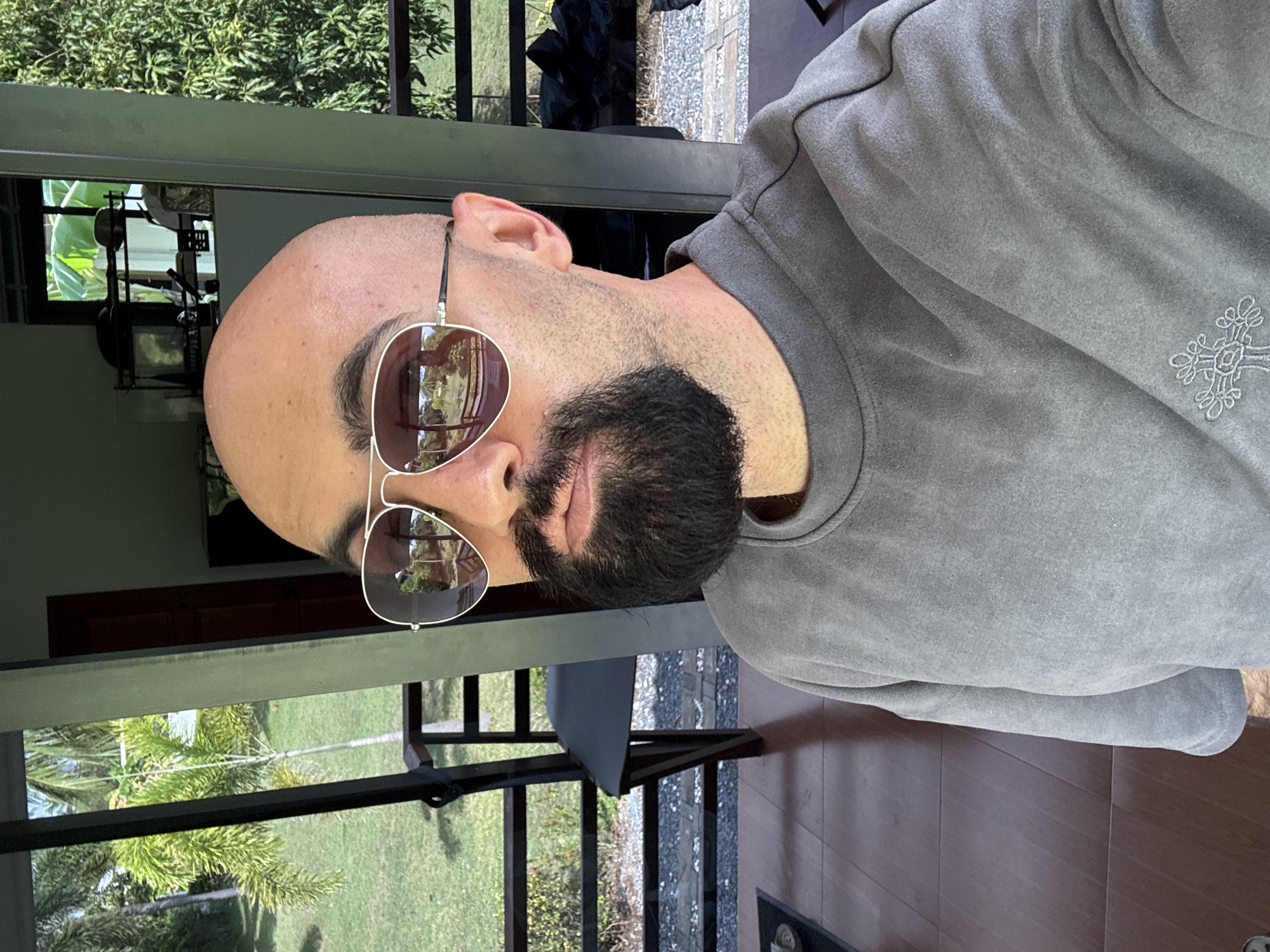 A bald man with a beard and sunglasses is taking a selfie. He is wearing a gray sweatshirt. The background shows a tropical outdoor setting with palm trees and greenery, likely a resort or private residence in Ko Samui District, Thailand. The man is standing on a tiled porch with a wooden floor.