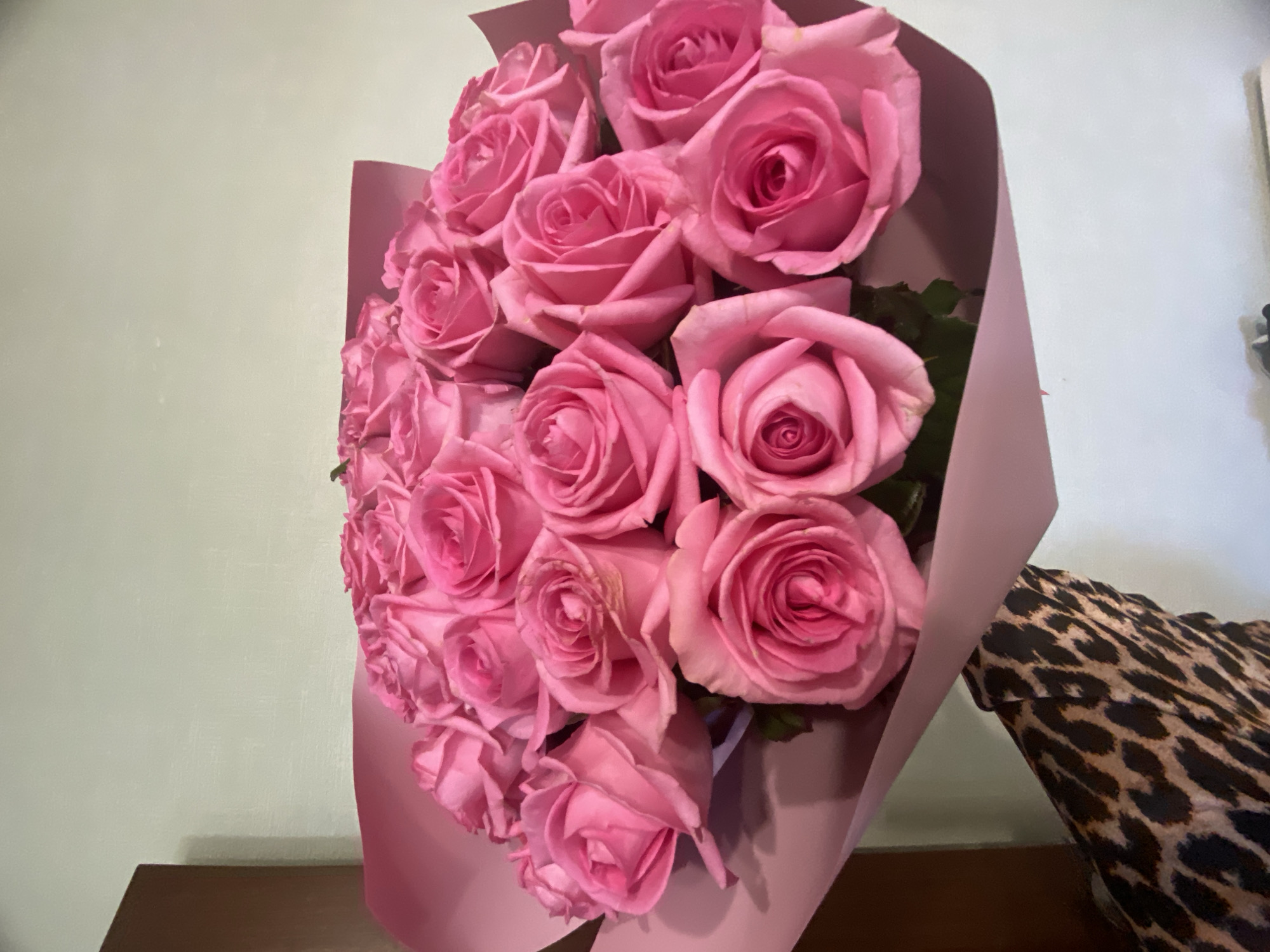 A bouquet of approximately 20 pink roses, wrapped in matching pink paper, is positioned on a brown surface against a pale wall. A portion of an object with a leopard print pattern is visible to the right of the bouquet. The location in the city of Sochi, Russia, is not discernible from the image.