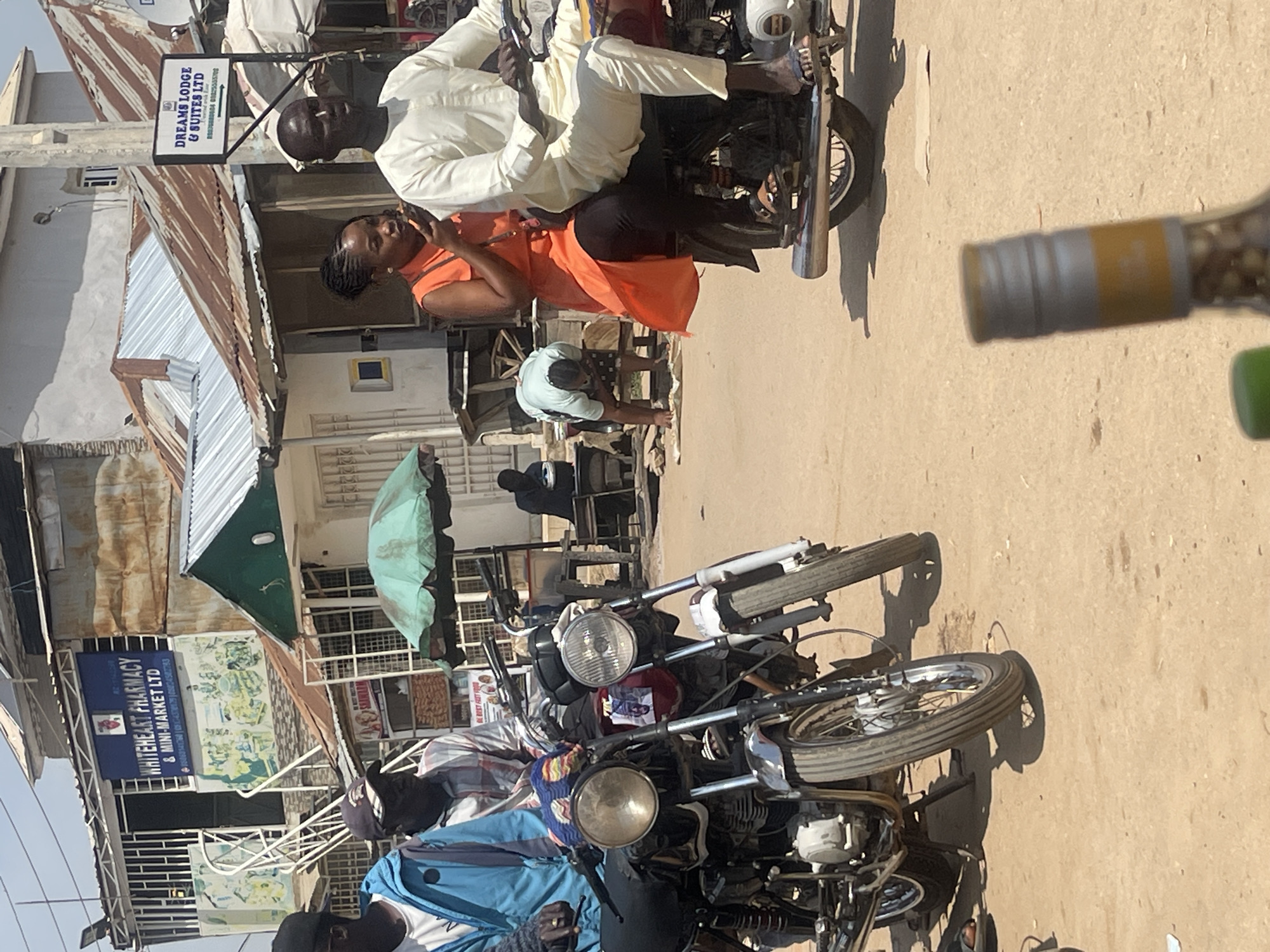 The image depicts a street scene in Abuja, Nigeria, likely in a commercial area. In the foreground, two motorcycles are parked side-by-side. The motorcycle on the left is black and maroon, while the one on the right is red and white. A man wearing a blue jacket and a dark cap is partially visible on the left. Next to him, another man in a checkered shirt and a baseball cap is also on a motorcycle.
On the right side of the image, a couple rides a motorcycle. The woman, wearing an orange dress and black leggings, is seated behind the man, who is dressed in a cream-colored outfit. They appear to be interacting.
In the background, buildings line the street. One building features a sign that reads "WHITEHEART PHARMACY & MINI-MARKET LTD". Another sign visible further down the street says "DREAMS LODGE & SUITES LTD". A green umbrella is set up near a stall. Several other individuals are visible in the background, including a person bending over and another person sitting at a table. The ground is covered in dirt and sand. The overall impression is a bustling urban environment.
