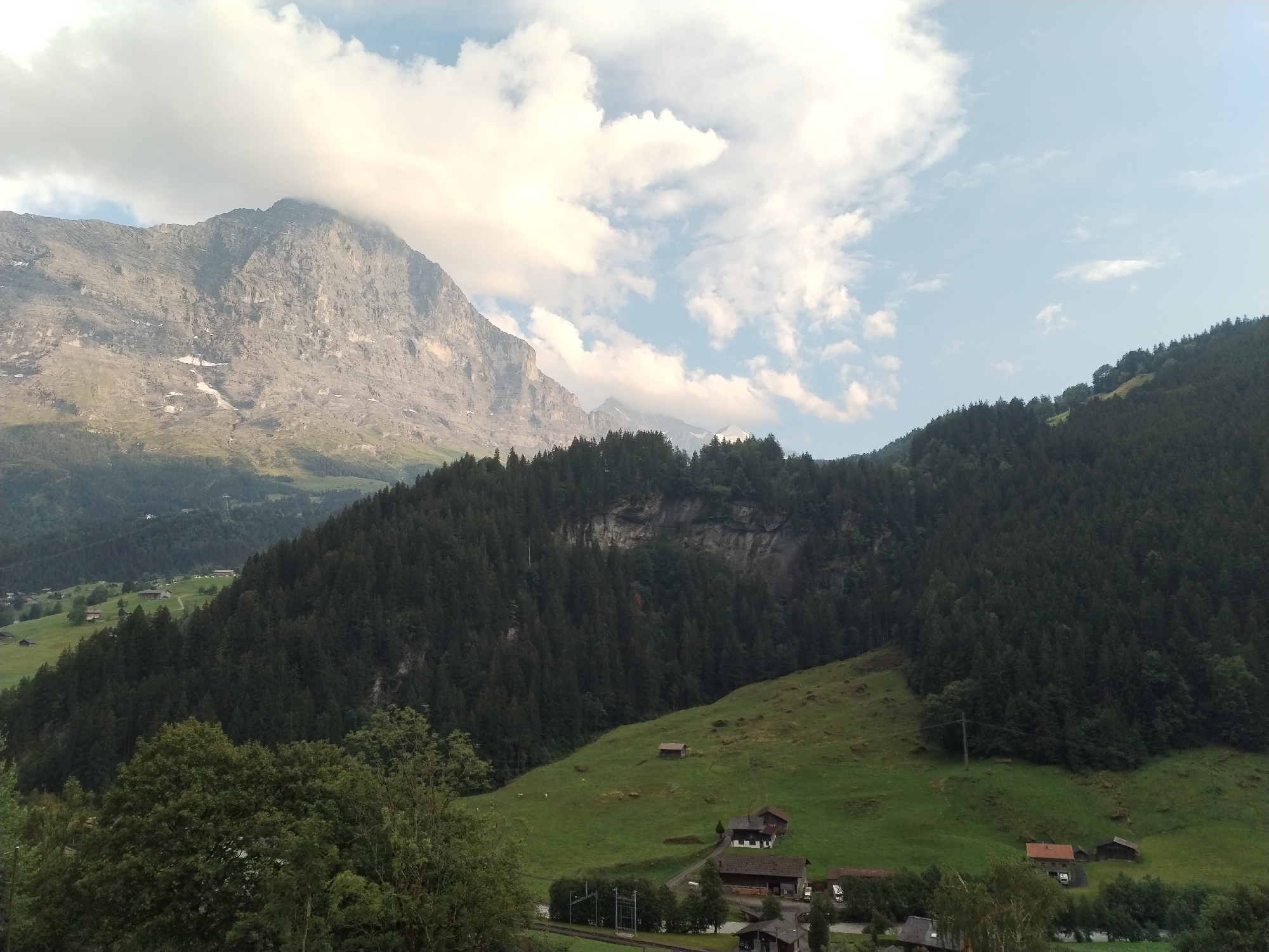 The image captures a vast, mountainous landscape with a mix of rugged peaks, dense forests, and verdant meadows. In the foreground, a lush green hillside is dotted with several rustic wooden buildings, including houses and smaller sheds, suggesting a rural settlement. A railway track with overhead power lines runs horizontally across the lower part of the frame, near a cluster of buildings. The middle ground is dominated by a steep slope covered in a thick, dark green coniferous forest, rising up towards a rocky mountainside. The imposing mountain range in the background is characterized by its weathered, grey rock faces, with patches of snow visible on its upper slopes. The sky above is a pale blue, interspersed with large, fluffy white clouds, some of which are casting shadows on the mountains. The overall impression is one of natural beauty and tranquility, characteristic of a mountainous region, likely in the Alps, given the architectural style of the buildings and the mountainous terrain. The scene appears to be captured during daylight, possibly late afternoon or early evening, due to the soft lighting and the angle of the sun.