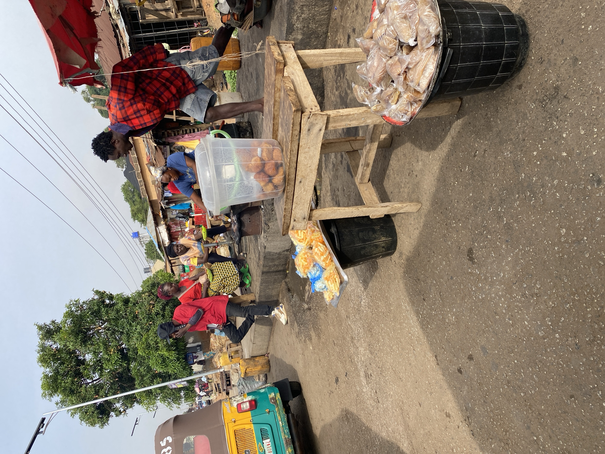 The image depicts a street scene in Bukuru, Nigeria. A man in a red plaid shirt and denim shorts stands at a wooden stall. The stall contains a clear plastic container filled with round fried foods, a black bucket filled with bags of bread, and additional bags of fried food hanging from the side. Another man in a blue shirt stands next to the stall. Further down the street, people are sitting and standing near what appear to be market stalls. A red-shirted man walks alongside a tricycle in the lower left corner of the image. A large tree is visible to the left of the scene. The street surface is asphalt. Overhead, power lines stretch across the frame.