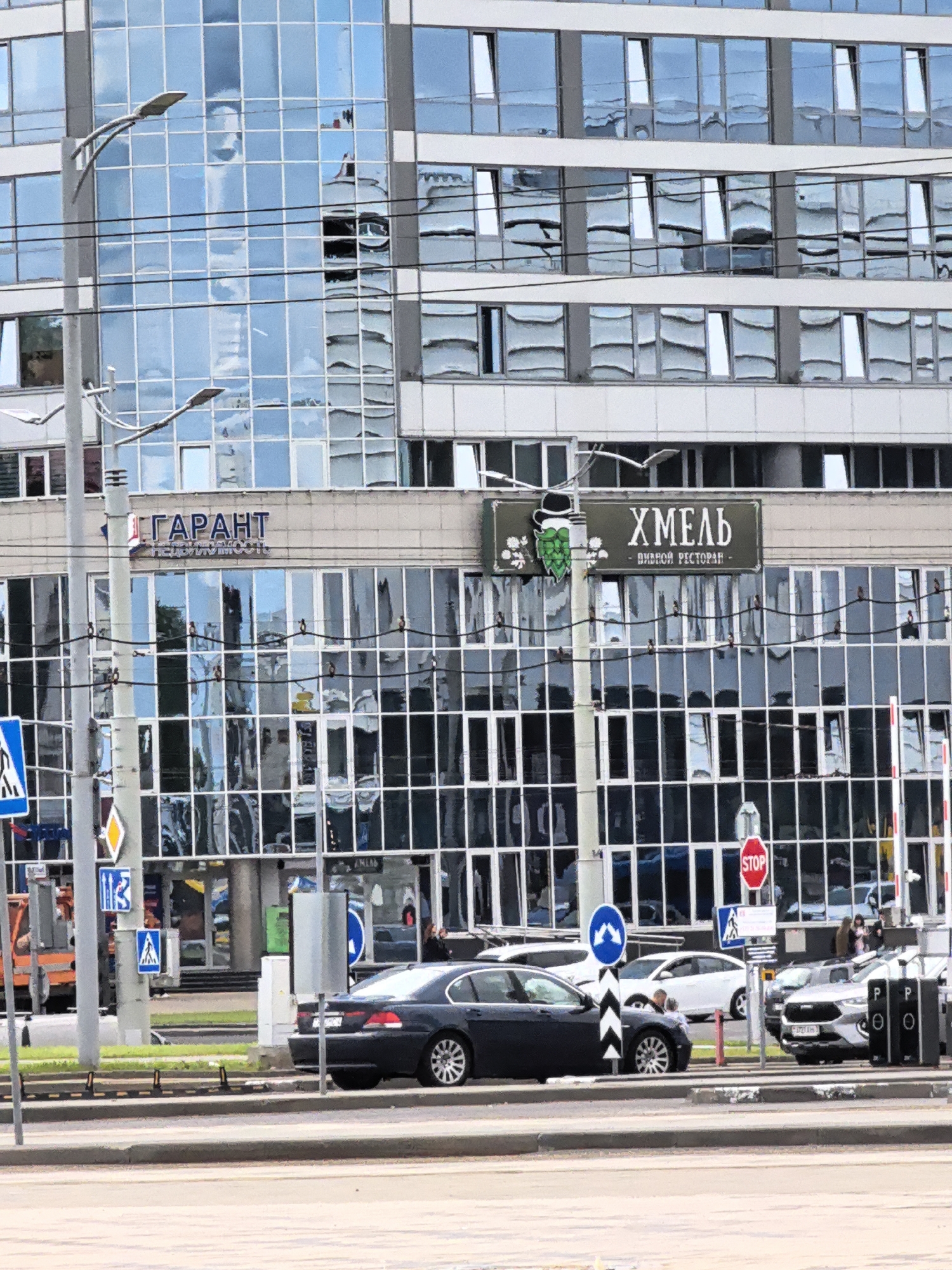 This is a medium shot, taken from across a road, of a modern office building with a glass facade. The building is several stories high, and the glass reflects the sky and surrounding environment, creating a distorted, abstract pattern. Two streetlights are visible in the foreground, attached to a grey pole.
On the building's facade, two signs are visible. The first, to the left, reads "ГАРАНТ" in large, bold, red letters, with smaller text below. To the right of that sign, a second sign, in a dark green rectangular frame, displays the word "ХМЕЛЬ" in large, white letters, with "ПИВНОЙ РЕСТОРАН" in smaller white letters below. The sign also features an illustration of a hop cone wearing a hat and a snowflake.
In the foreground, a dark-colored sedan is parked on the side of the road, facing left. Several other cars are visible behind it. Traffic signs are also present, including a blue and white pedestrian crossing sign, a yellow diamond-shaped sign, and a red octagonal stop sign. A construction vehicle, orange and grey, is partially visible on the far left of the image.
The overall scene appears to be an urban environment. The lighting suggests it is daytime, and the reflections on the glass indicate a bright sky, possibly with some clouds. There are no clear indications of specific weather conditions beyond general brightness. There are a few people visible in the background on the sidewalk near the building, but they are too small and indistinct to discern any specific activity or emotion. The presence of traffic, buildings, and signage points to a busy commercial or business district.