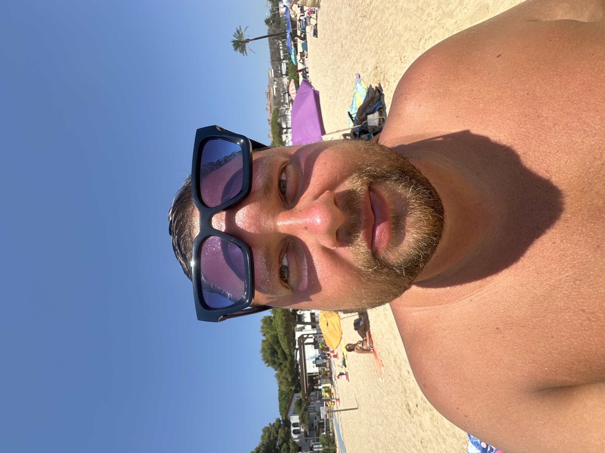 The image is a selfie taken on a sunny day at a beach in Sant Feliu de Guíxols, Spain. A man with sunglasses perched on his forehead is in the foreground, his face and upper torso filling most of the frame. He has light skin and a beard. The background shows a sandy beach with several people relaxing under umbrellas, some sunbathing and others sitting in chairs. There are trees and buildings visible in the distance, along with palm trees. The sky is clear and blue. The scene depicts a typical day at a coastal resort.