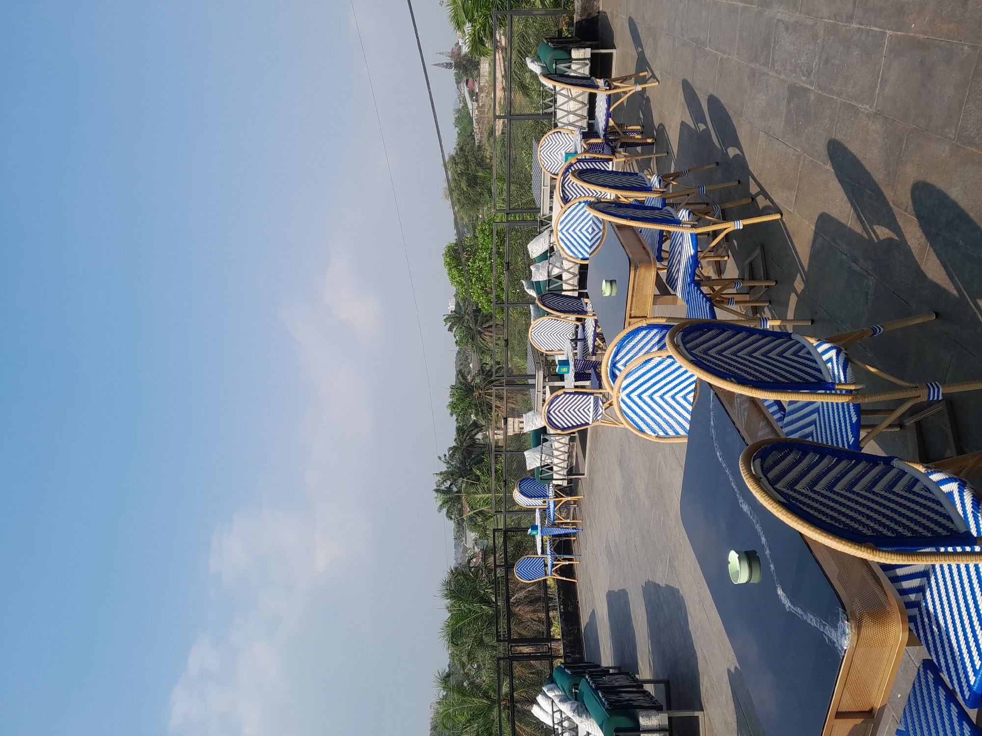 The image depicts an outdoor seating area of a cafe or restaurant. The foreground features several tables with dark tabletops, some with chairs arranged around them. The chairs have a distinctive woven rattan frame and blue and white striped upholstery. An ashtray sits on one of the tables. Beyond these tables, more seating arrangements are visible, including lounge-style seating with plush cushions. A black metal railing encloses the seating area, beyond which a verdant landscape with palm trees and other foliage is visible. In the distance, buildings and structures suggest an urban or semi-urban setting. The sky above is a clear, bright blue with a few scattered white clouds. The overall impression is one of a relaxed, open-air dining or lounging space. The location is identified as Sempusari, Indonesia.