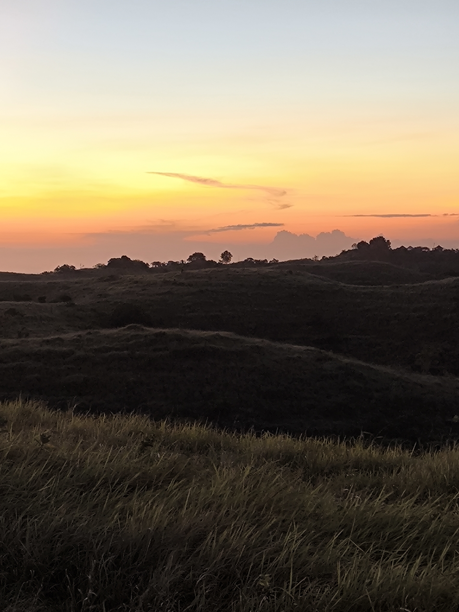 The image captures a sunset over a grassy landscape in Nusa Penida, Indonesia. The sky transitions from light blue at the top to orange and yellow near the horizon, where a thin cloud formation is visible. In the middle ground, a series of rolling hills are silhouetted against the sunset, dotted with trees. The foreground is filled with tall grass. The scene appears serene and uninhabited.