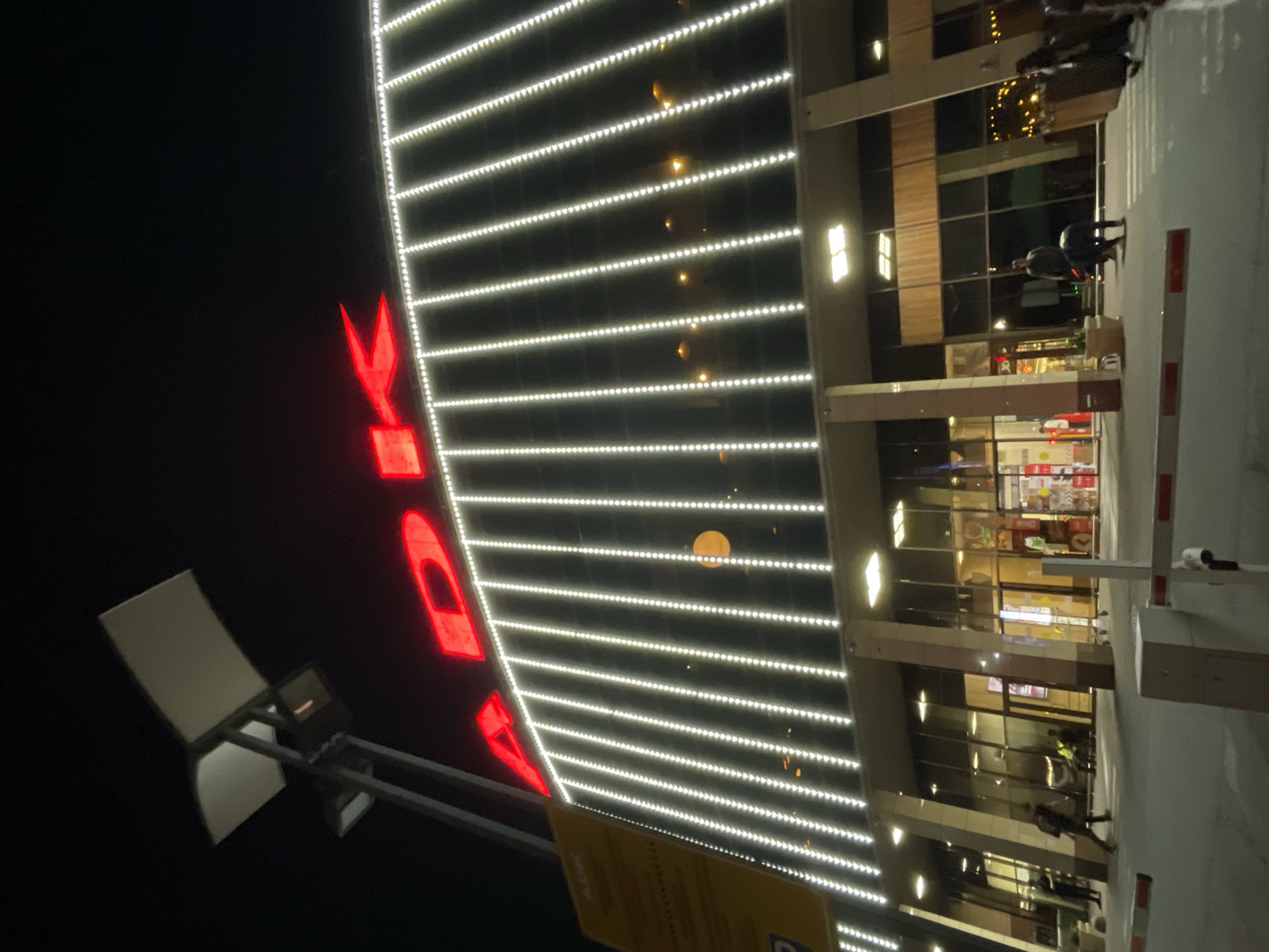 The image shows a nighttime exterior view of a large building with prominent red neon lettering that reads "ADK". The building's facade is decorated with vertical rows of white LED lights. In the foreground, a pedestrian crossing with a red and white barrier is visible. Several people are seen walking or standing near the entrance of the building. The building appears to be a shopping mall or entertainment complex, likely located in Almaty, Kazakhstan, given the "ADK" signage which is associated with the ADK Mall in Almaty. The sky is dark, indicating it is nighttime.