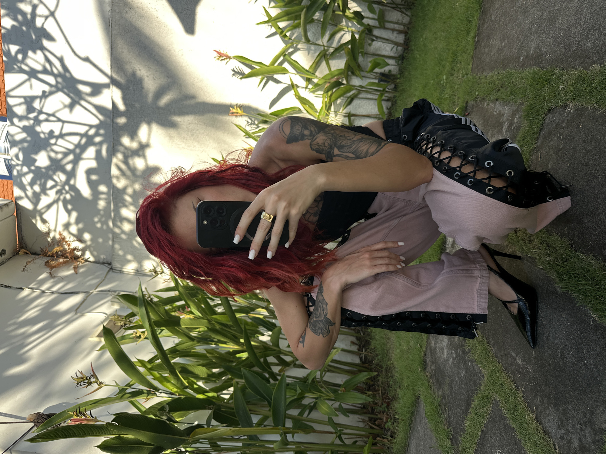 A woman with bright red, wavy hair crouches outdoors, holding a smartphone to take a selfie. She is wearing a black strapless top, light pink pants with black lace-up detailing along the sides, and black pointed-toe high-heeled shoes. She has tattoos on her arms. She is positioned on a paved area surrounded by grass, with lush green plants and a white wall in the background. Sunlight casts dappled shadows from foliage onto the wall. The location is North Kuta, Indonesia.