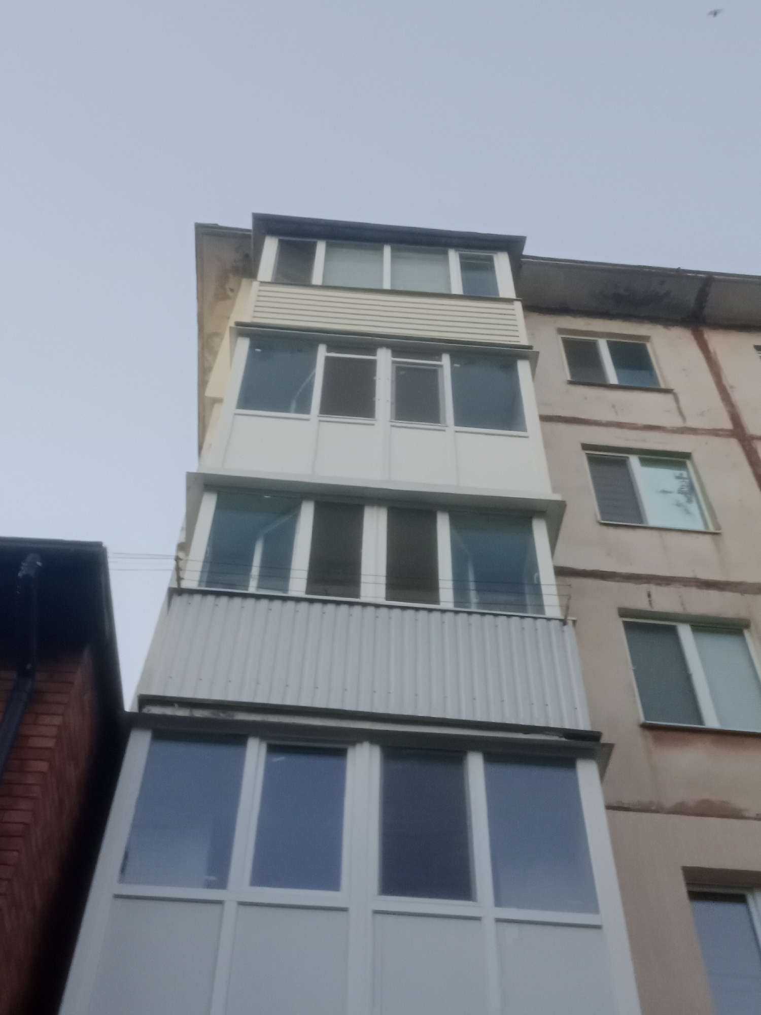 An exterior, low-angle, eye-level shot captures the side of a multi-story apartment building and an adjacent, narrower brick structure. The sky is overcast and light gray. The apartment building features a vertical addition to its facade, comprised of enclosed balconies that extend upwards across multiple floors. These balconies have white frames and windows, with the lowest section featuring corrugated white siding. The main facade of the apartment building is a muted beige color with visible horizontal lines and some signs of wear and discoloration. Windows on the apartment building are rectangular with white frames. The adjacent structure to the left is made of red brick and has a dark roofline. The scene is typical of an urban residential area in Horishni Plavni, Ukraine.