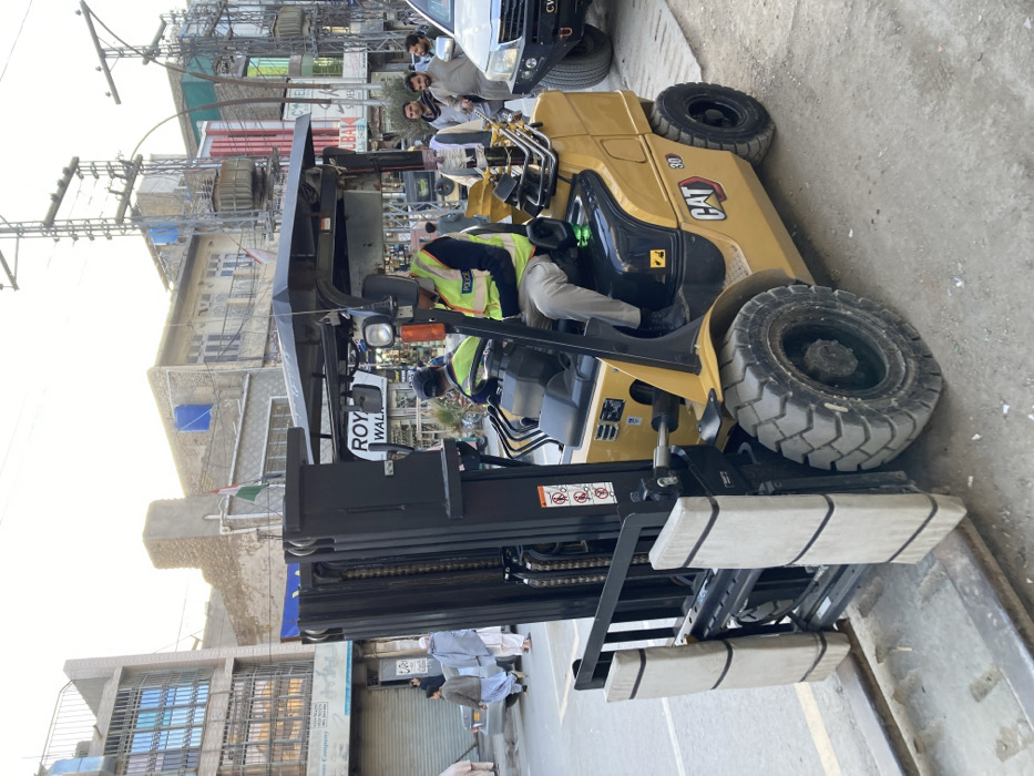 The image captures an urban street scene in Quetta City Tehsil, Pakistan, under daylight conditions. In the foreground, a yellow CAT 30 forklift is stationary, positioned on what appears to be a concrete curb or platform adjacent to a paved road. The forklift's front mast is black, and its forks are equipped with light-colored protective padding. A male operator, dressed in a light-colored shirt, grey trousers, and a green high-visibility vest marked "POLICE," is seated in the driver's seat, appearing to look down at his lap or hands. A second individual, also in a green high-visibility vest, is partially visible behind the primary operator.
To the right, a white Toyota Hilux-type SUV, fitted with an aftermarket steel front bumper, is parked on the street. The background features multiple multi-story commercial and residential buildings constructed from brick and concrete, some displaying visible power infrastructure including overhead lines and transformers on utility poles. A blue water tank is visible on the roof of one building, and a small green and white flag is draped on another facade. Signboards are present, with one clearly displaying "ROYAL WALKS" and another partially visible for "LUBAI SHOES." Several male pedestrians, attired in traditional shalwar kameez, are observed walking and standing on the street and near the buildings. The ground in the immediate foreground consists of dirt and scattered debris, transitioning to a paved road surface.