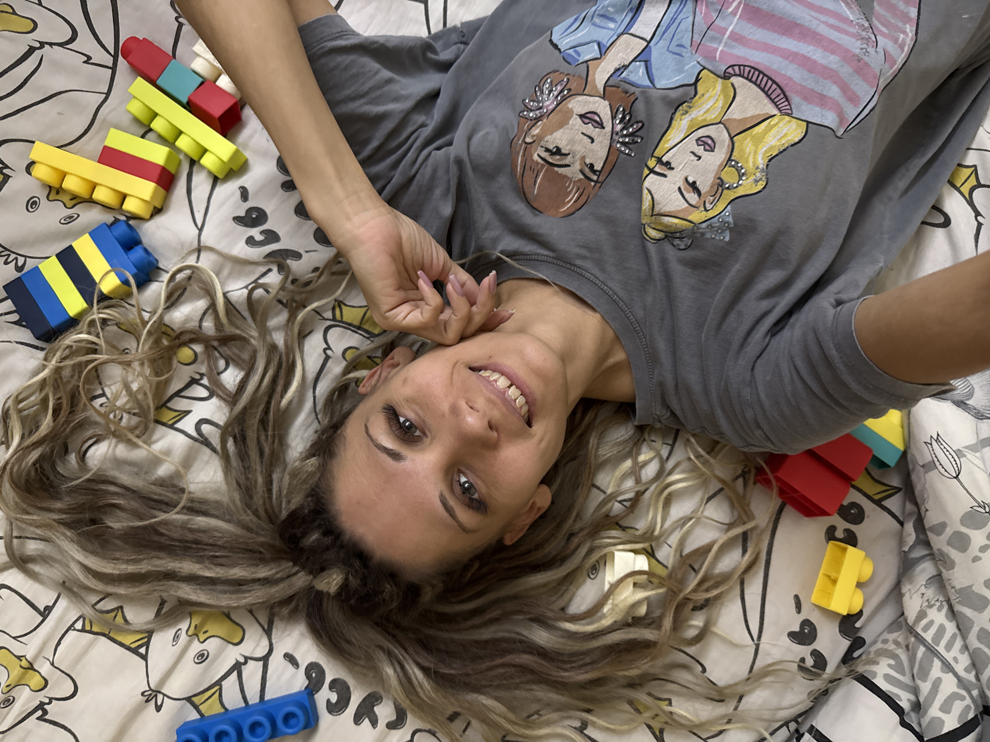 In Rusanovo, Russia, a woman is lying on her back on a white bedsheet with a cartoon duck pattern. She has long, wavy, blonde and brown hair spread around her head. She is wearing a grey t-shirt with a graphic print of two cartoon women. Her left arm is bent at the elbow and resting near her head, while her right arm is extended along her side. In the background, scattered on the bedsheet, are colorful building blocks in blue, yellow, red, and green.