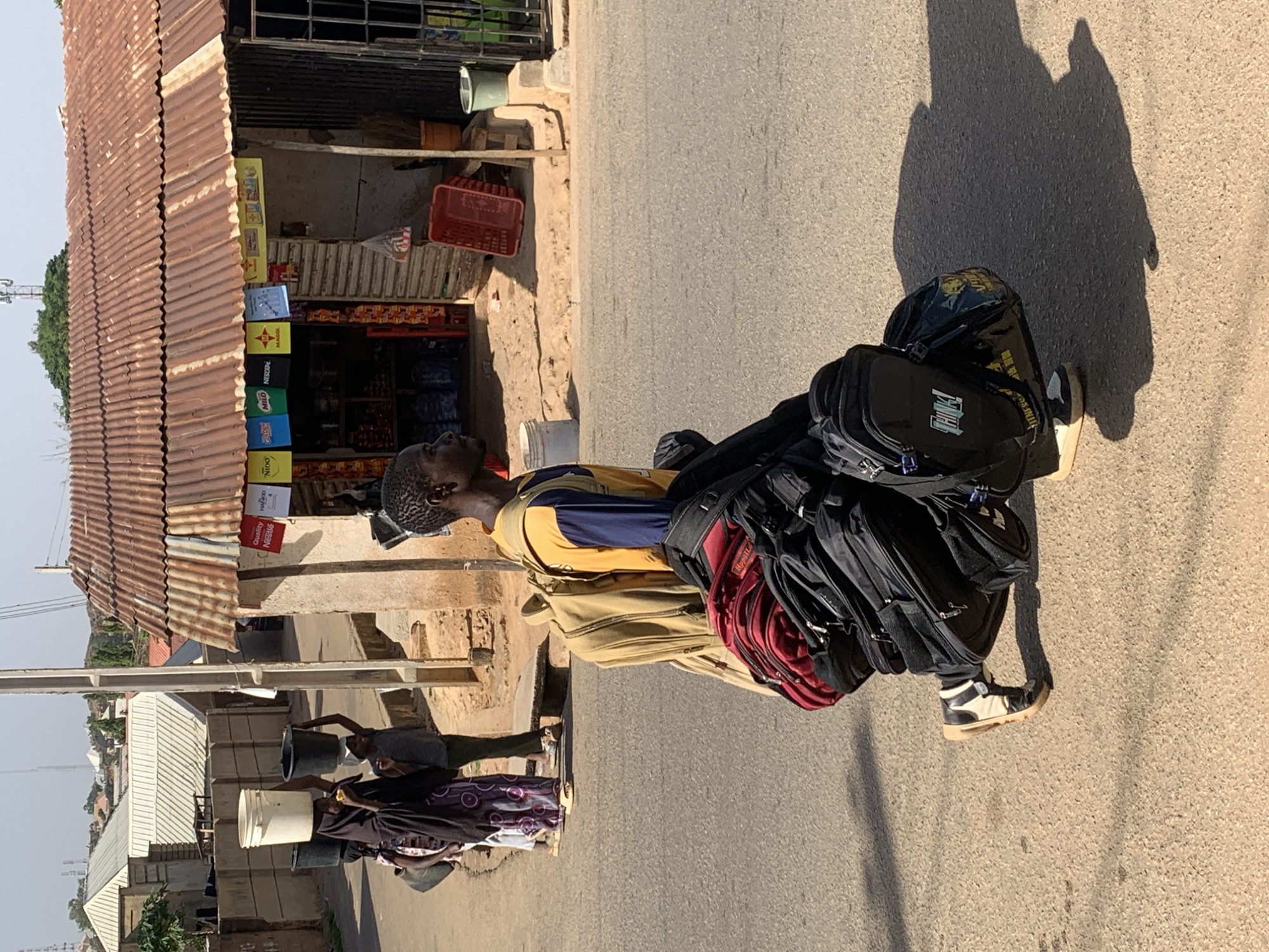 The image captures a street scene in Jos, Nigeria. A man stands on a paved road, carrying a large stack of backpacks. He wears a yellow and blue shirt, khaki pants, and black and white high-top sneakers. Several black backpacks are stacked on his back, with a red one visible in the middle. His shadow stretches onto the road next to him. In the background, a roadside shop with a corrugated metal roof displays various goods and products. Further down the road, two people are carrying buckets on their heads. Utility poles and residential buildings are also visible. The sky is pale, suggesting daylight.