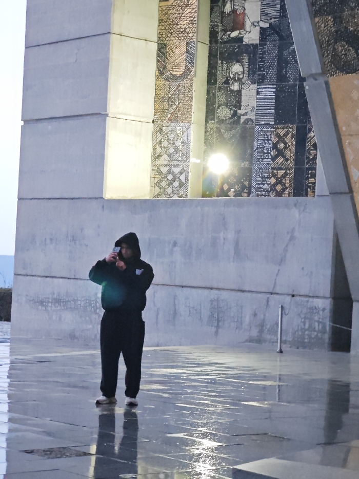 The scene depicts a male individual standing on a wet, paved plaza in front of the Monument to 1300 Years of Bulgaria (Founders of the Bulgarian State Monument) in Shumen, Bulgaria. The colossal concrete and granite monument features internal mosaic murals with depictions of historical figures and abstract geometric patterns, illuminated by a distinct bright light source visible within the structure. The individual, dressed in a dark hooded sweatshirt with the hood up, dark trousers, and light-colored athletic shoes, holds a smartphone-like device with both hands at chest level, angled towards the monument, indicating the action of photographing or video recording. The ground is reflective due to moisture, showing clear reflections of the individual and the bright light. A slender metallic post or bollard is visible on the pavement to the right. A distant, indistinct landform is visible on the far-left horizon.