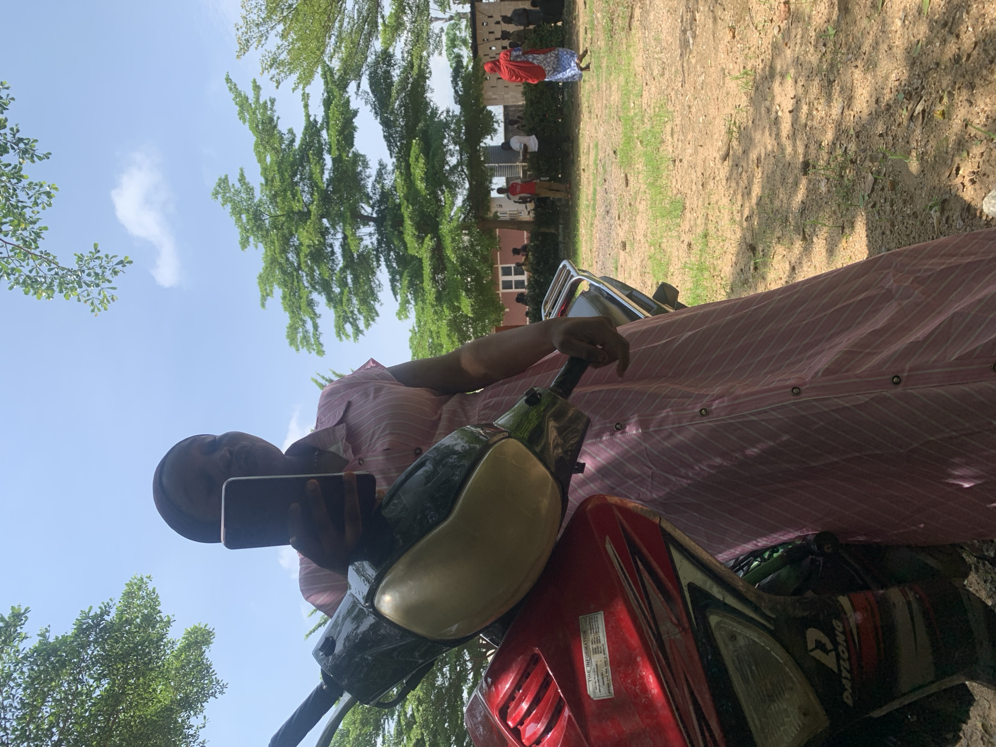 A medium shot captures a woman, seen from the waist up, wearing a pink and white striped dress and a dark headscarf. She is holding a dark smartphone in her right hand, appearing to take a selfie or record a video. The woman is positioned in front of a red and black motorcycle, with the handlebars and front of the vehicle prominently in the foreground. The background reveals a sunlit outdoor environment with a clear blue sky dotted with fluffy white clouds. Several trees with lush green foliage are visible, some casting shadows on the ground. In the middle ground, a group of people are gathered near a building with white window frames, and a woman in a red hijab and blue patterned dress is walking away from the camera towards the right. The ground is a mix of dry dirt and sparse green grass. The scene appears to be in a public space, possibly a roadside or a communal gathering area, in Akwanga, Nigeria.