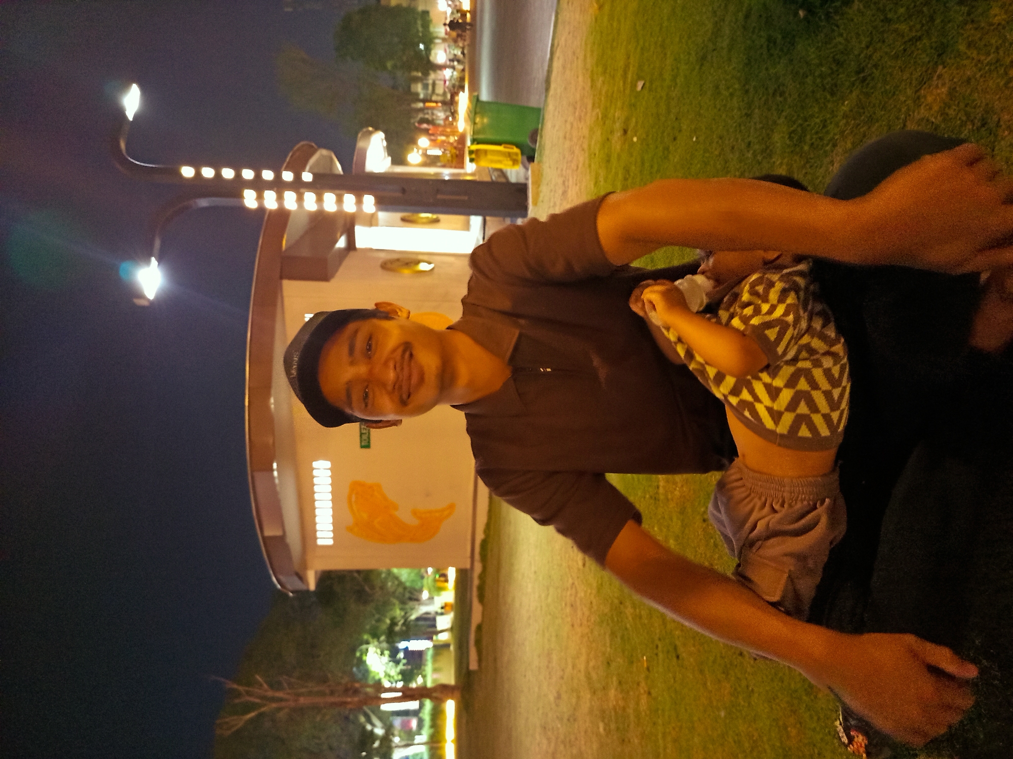 In Sidoarjo, Indonesia, at night, a man wearing a brown polo shirt and a dark baseball cap is seated outdoors on a grassy area. He is smiling at the camera and has a mustache. A young child, partially visible and wearing patterned clothing, is being held on his lap. The background features a white circular building with a yellow fish mural and a street lamp with multiple lights. The area appears to be a public park or recreational space.