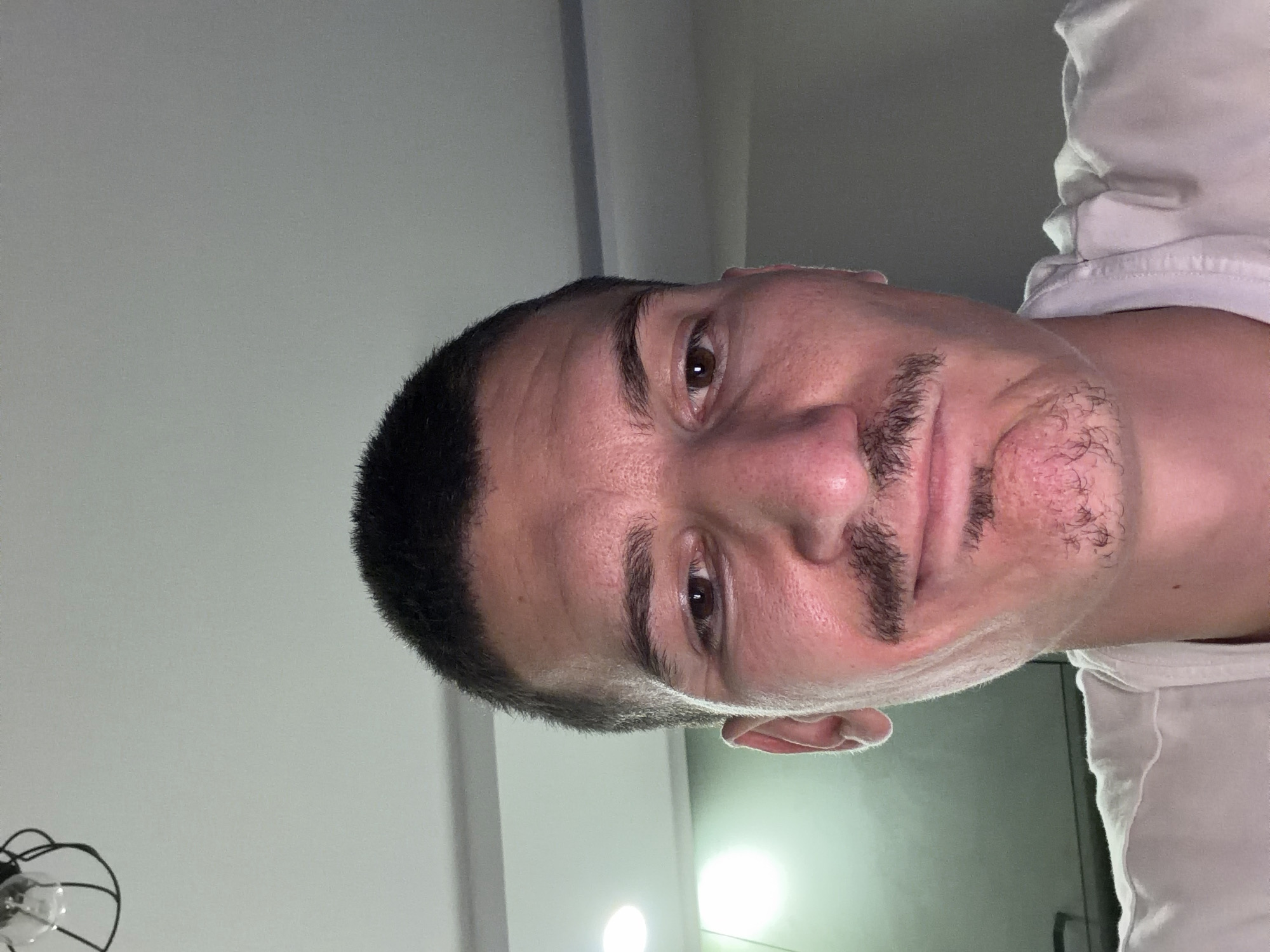 A close-up, low-angle shot of a man's face, tilted slightly to the right. He has dark hair, brown eyes, and a mustache. He is wearing a white shirt. The background consists of a white wall and some lighting fixtures. The location is not discernible from the image, so I am unable to confirm Sofia, Bulgaria.