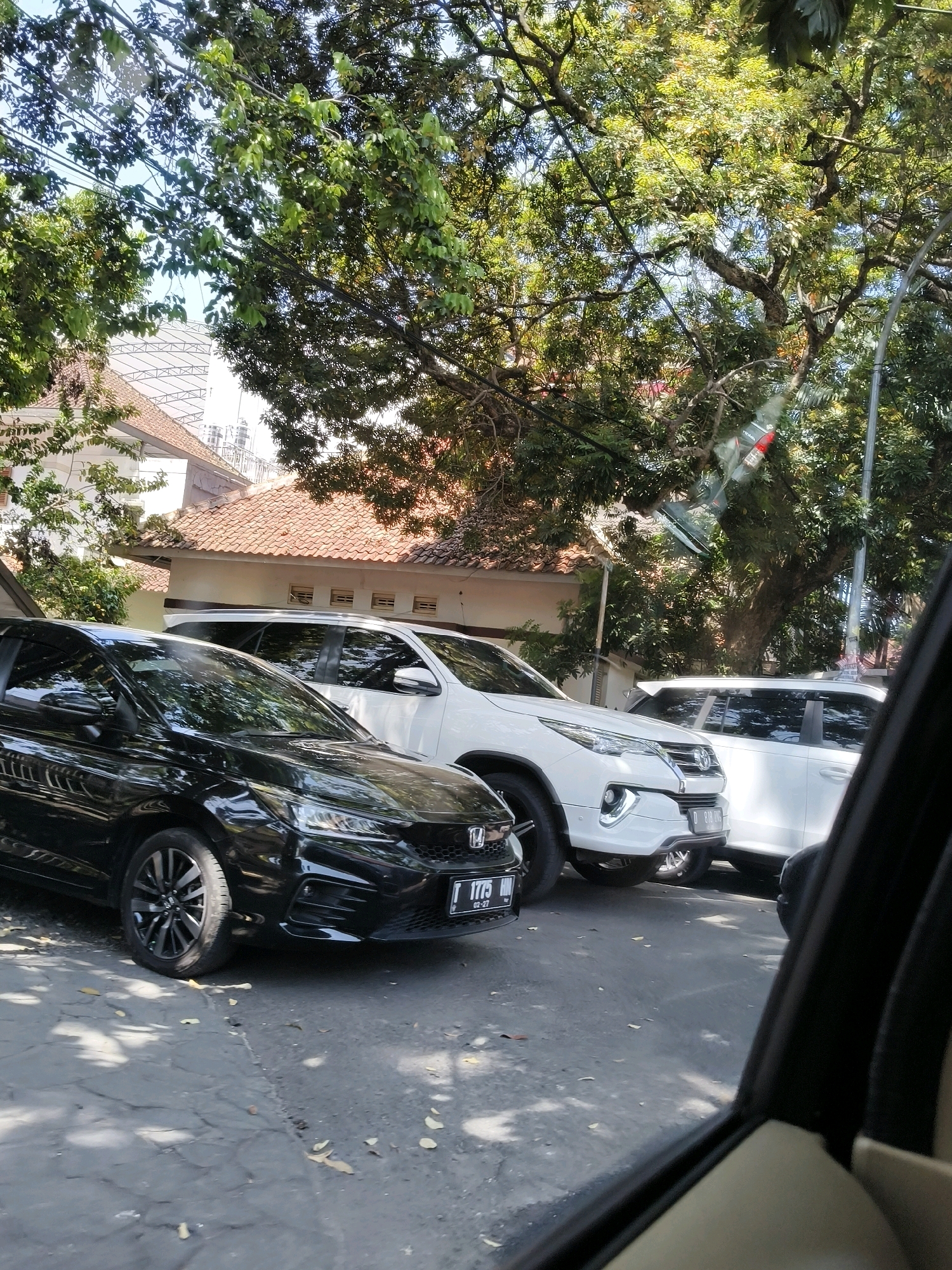The image depicts a street scene in Bandung City, Indonesia, characterized by parked vehicles and lush greenery. In the foreground, a black sedan is prominently displayed, its front end angled towards the viewer. Behind it, a white SUV is parked, with another white vehicle partially visible to its right. The vehicles are situated on a paved road, with fallen leaves scattered across the surface. Above the cars, the branches of large trees laden with green foliage create a canopy, filtering the sunlight and casting dappled shadows on the ground. In the background, a building with a terracotta-tiled roof and white walls can be seen, hinting at residential or commercial structures within the urban environment. The overall impression is one of a sun-drenched day in a leafy urban setting.