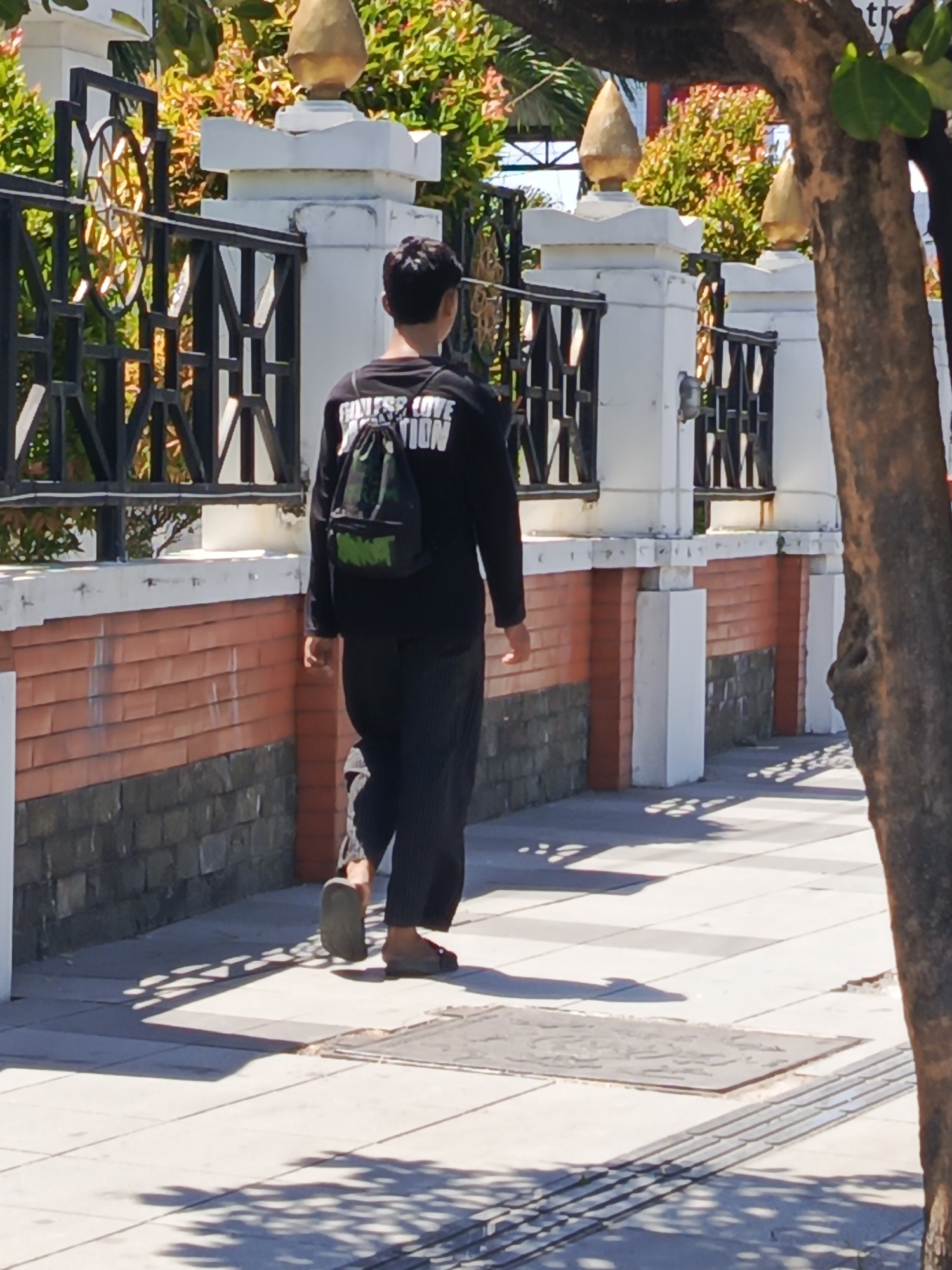 A young man walks away from the camera on a sunlit sidewalk in Surabaya, Indonesia. He is wearing a black long-sleeved shirt with white text and a small black and green backpack. His pants are dark and baggy. He is wearing open-toed sandals. The sidewalk is paved with light-colored tiles and has a textured tactile paving strip along the edge. Sunlight casts dappled shadows on the sidewalk, suggesting there are trees overhead. To the left, a low brick wall with stone detailing borders the sidewalk. Behind the wall is a black metal fence with ornate detailing and white pillars topped with golden finials. Lush green and orange foliage is visible behind the fence. A thick tree trunk is on the right side of the frame, also casting shadows.