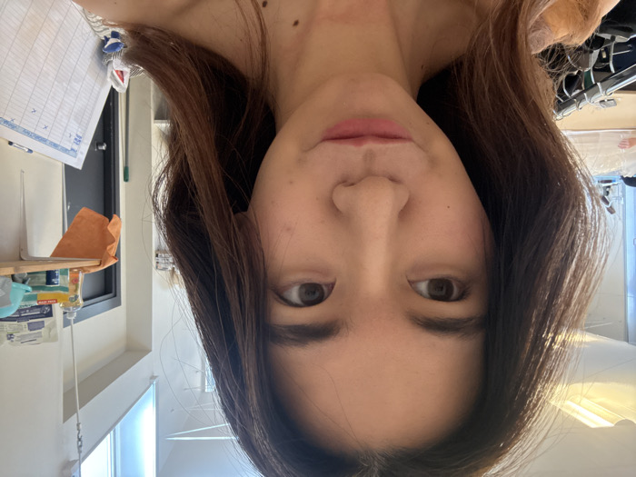 The image presents a close-up, selfie-style photograph of a young East Asian female in an indoor environment situated in Arcueil, France. The female subject has long, dark brown hair and light brown eyes, looking towards the right side of the frame with her lips slightly pursed. Moles are visible on her right neck and upper chest area. She is wearing an off-the-shoulder garment, appearing light peach or nude in color.
The background on the right features a light-colored wall with a partially visible dark door. A light wooden shelf is mounted on this wall, holding a yellow plastic bottle, a blue container, a smaller yellow container, and a light blue hard hat. An orange cloth is draped over the shelf's front edge. Below the shelf, a white grid board, marked with "X" in several cells, leans against the wall, next to a visible green mop or broom handle. Above the shelf and door, a high window or skylight is present, and a metal rod is suspended from the ceiling.
The background on the left is blurred but shows overhead fluorescent lighting fixtures, a clothing rack with light-colored garments on hangers, and a section of a workbench or table.