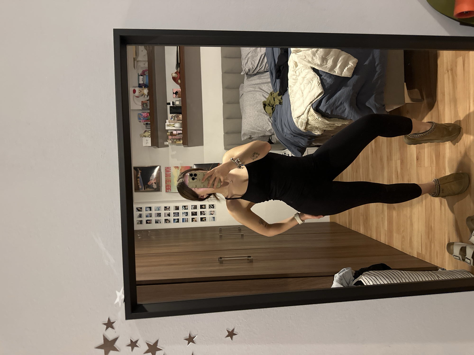 A person stands in a bedroom, taking a mirror selfie. The bedroom contains a bed with multiple pillows and blankets, a shelf with various items, and a wooden cabinet. The person is wearing a black jumpsuit and slippers. Star-shaped decorations are on the wall next to the mirror. The location is an indoor space in Huixquilucan de Degollado, Mexico.