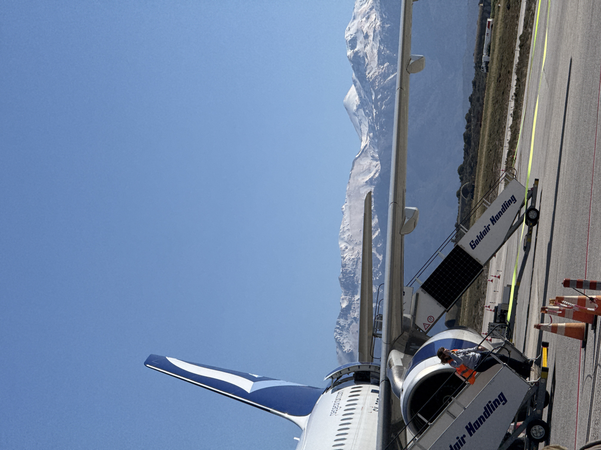 The image captures an airplane parked on the tarmac at Chania International Airport in Kounoupidiana, Greece. The aircraft is fitted with mobile stairs labeled "Goldair Handling," and an individual wearing a gray shirt and orange safety vest is visible on the stairs, seemingly inspecting the aircraft. The sky is a clear blue, and in the distance, a snow-capped mountain range is visible, likely the Lefka Ori (White Mountains) of Crete. The ground is marked with yellow and red lines, standard markings for airport taxiways and aircraft stands. Several orange and white cones are placed near the stairs.