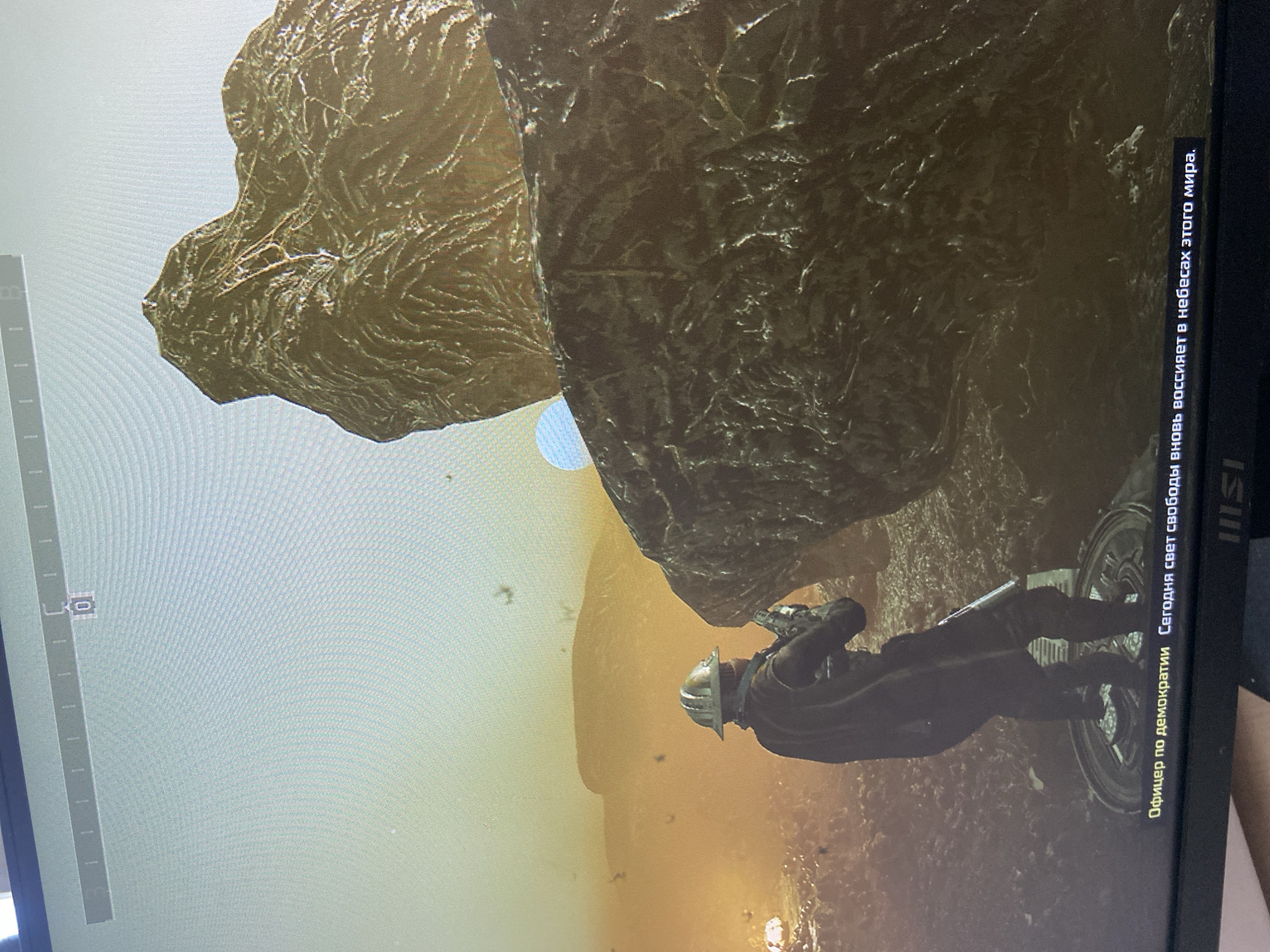 The image shows a video game scene with a person in a helmet and protective gear holding a weapon, standing on a platform in a rocky, alien-like environment. In the background, there are desolate landscapes with large rock formations and a hazy, orange sky. A small, pale blue orb is visible in the sky. Text in Russian appears at the bottom of the screen. The scene does not depict any location in the city of Beloretsk, Russia.