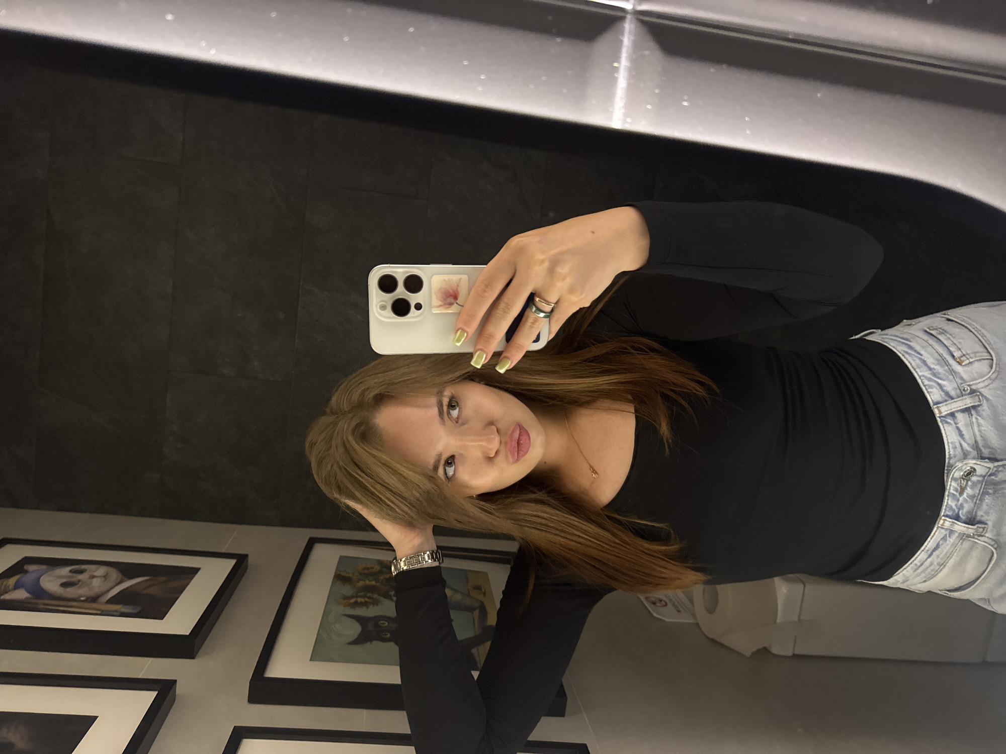 A woman is taking a mirror selfie in a restroom. She has long, light brown hair and is wearing a black long-sleeved shirt and light-wash jeans. She is holding her phone in her right hand, which is angled towards the mirror. Her left arm is raised, with her hand resting on her head. She is wearing rings on her right hand and a watch on her left wrist. The background shows a dark tiled wall, framed pictures on the wall, and a toilet with a roll of toilet paper. The location is in the city of Chaikovsky, Russia.