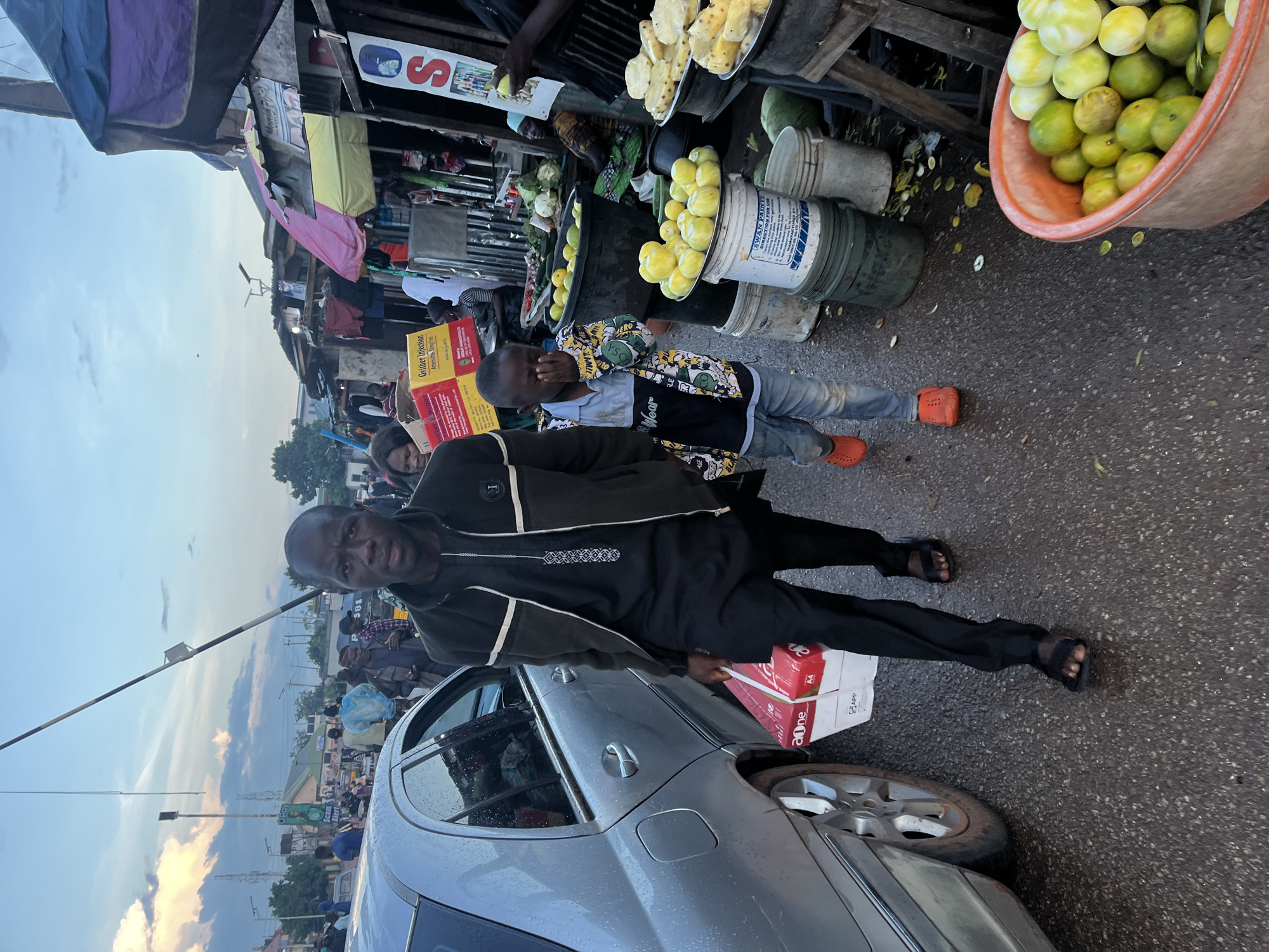 The image depicts a street scene in Gura Top, Nigeria, likely during twilight. A man in a dark jacket and black traditional attire walks towards the viewer, carrying a red and white box of paper in his left hand. Beside him, a young boy in a colorful shirt and jeans walks with his right hand covering his eyes, wearing orange clogs. To their left is the side of a silver car. The background shows a busy marketplace with various stalls, some covered with tarps, selling goods including fruits. Several people are visible in the background, suggesting a bustling atmosphere. The sky is cloudy with hints of sunset colors. A white pole with a cable stretches across the upper part of the frame.