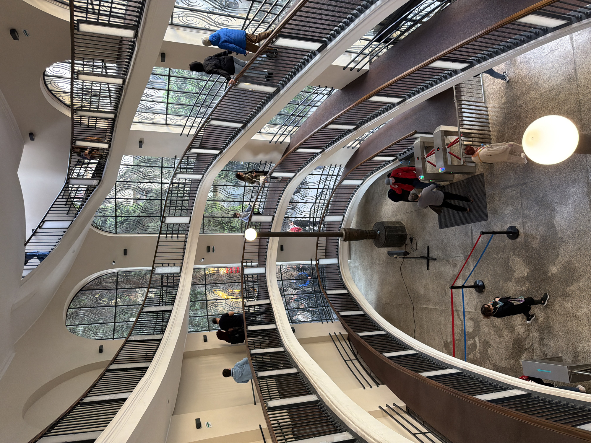 The image is an interior view of the Bank of Georgia headquarters building in Tbilisi, Georgia, designed by architects George Chakhava and Zurab Jalaghania. The perspective is from above, looking down into the atrium. The building features a series of stacked, staggered volumes. The atrium is characterized by multiple floors connected by walkways with dark brown railings and open stairs. Large arched windows are visible on the exterior walls, providing natural light. People are seen on various levels, some walking, some standing. At the ground level, there are turnstiles and a light fixture. A red and blue structure is also visible on the floor, along with various pieces of equipment attached to the wall. The overall impression is a modern architectural space with multiple levels and an open, airy feel.
