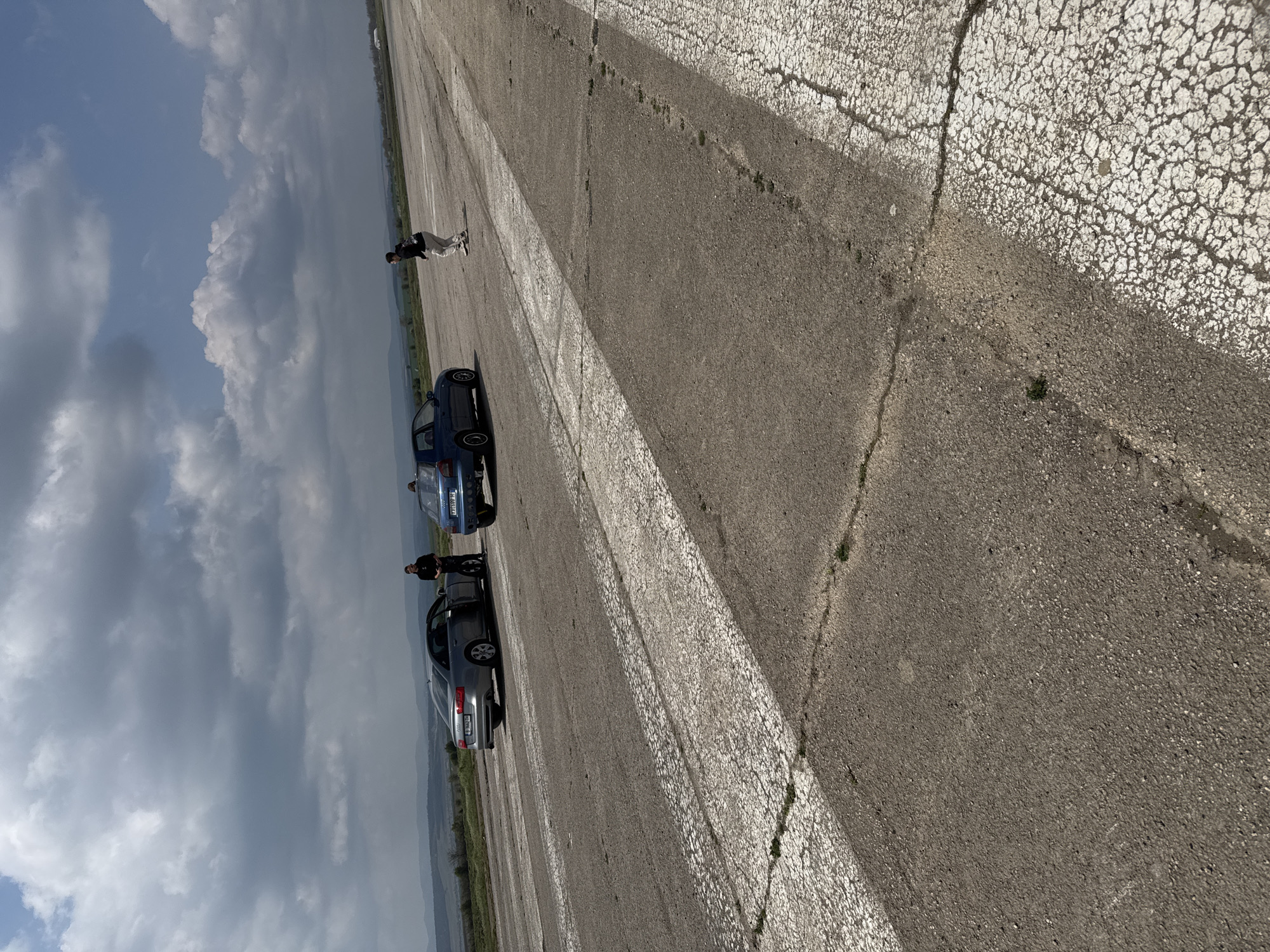 The image shows a cracked concrete surface, possibly a road or runway, in Buhovtsi, Bulgaria. Two cars, one blue and one silver, are parked on the left side. Two people stand near the silver car. A third person is standing at a distance away from the cars. The sky is cloudy.