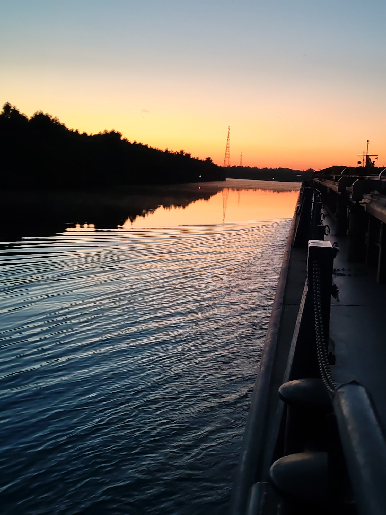 The image depicts a tranquil water scene at dusk, likely taken from the deck of a vessel. The sky transitions from a pale blue at the top to a warm orange and yellow gradient near the horizon, suggesting either sunrise or sunset. Silhouetted trees line the left bank of a calm body of water, reflecting the colors of the sky. In the distance, a tall, lattice-like tower, possibly a communication mast, is visible against the horizon, along with a smaller structure of a similar kind. On the right side of the image, the perspective is dominated by the side of a dark-colored vessel, revealing metal railings, bollards, and a heavy chain. The water's surface is marked by gentle ripples, creating a textured pattern that stretches towards the horizon. The overall impression is one of peacefulness and solitude in a natural, yet somewhat industrialized, setting. The specific location in Annensky Most, Russia, would likely be on the Svir River, a major waterway that flows through the region and is known for its barge traffic and associated infrastructure.
