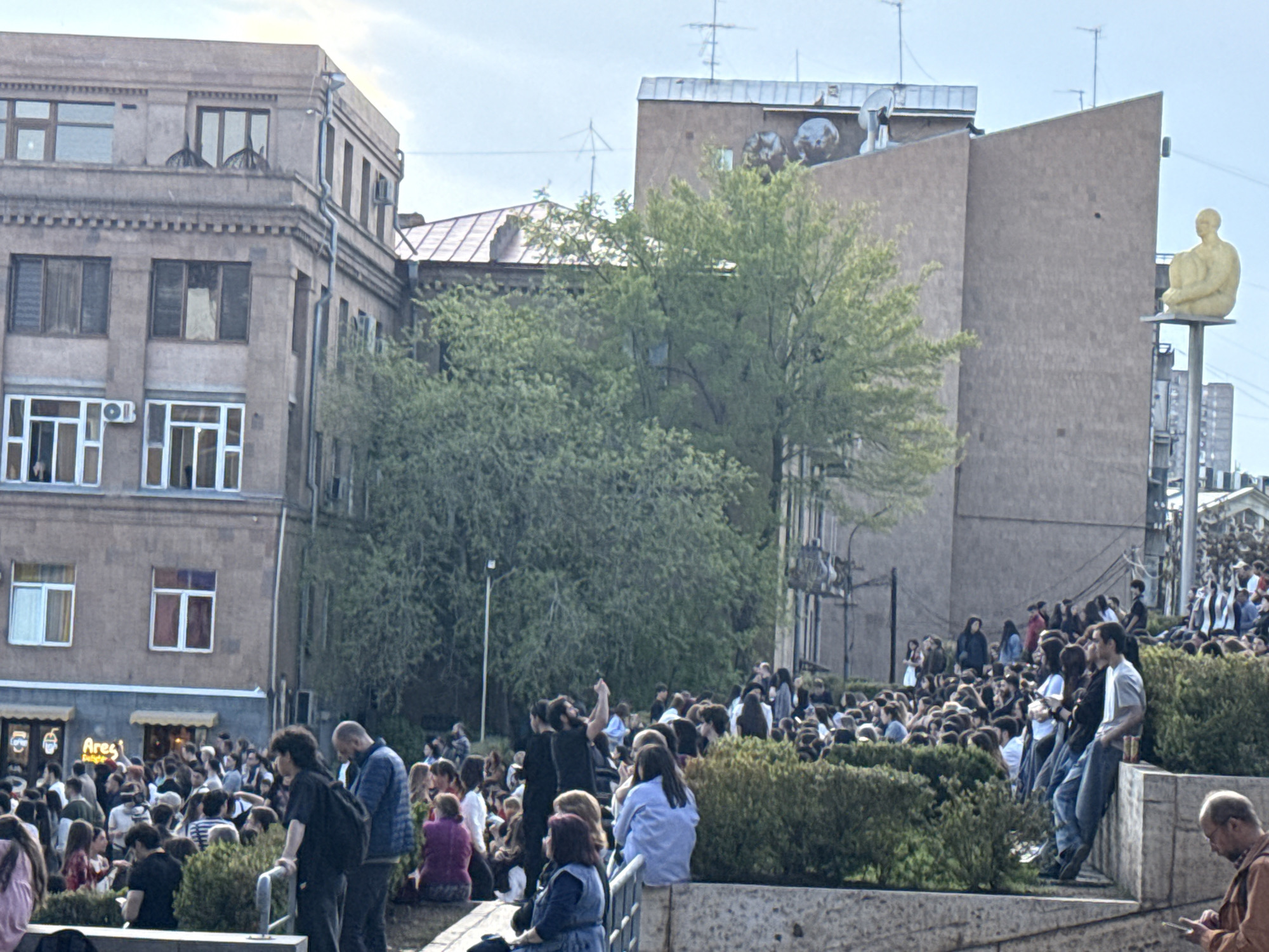 The image depicts a lively outdoor scene in an urban environment, identified as Yerevan, Armenia. A significant crowd of people, largely composed of young adults, fills the foreground and mid-ground. They are engaged in various activities: standing, conversing, and some are seated on low walls or ledges. One person in the crowd can be seen with an arm raised, possibly gesturing or cheering.
The background features several buildings characteristic of an urban setting. On the left, a multi-story building with a brown facade, multiple windows, and decorative cornices is visible. On the right, another building with a more modern, angular design stands. Perched high on a pole, against the backdrop of this building, is a prominent golden, minimalist statue of a seated human figure, known as "The Observer" or "Man" by Jaume Plensa. Rooftops are adorned with antennas and satellite dishes.
Green trees and bushes are scattered throughout the scene, suggesting landscaped public spaces. The bright sky indicates it is daytime, likely late afternoon or early evening, with clear weather suitable for outdoor gatherings. On the ground level of one of the buildings, a yellow awning displays the text "Ares Burger". The overall impression is of a bustling city center, vibrant with public life and distinct architectural and artistic landmarks.