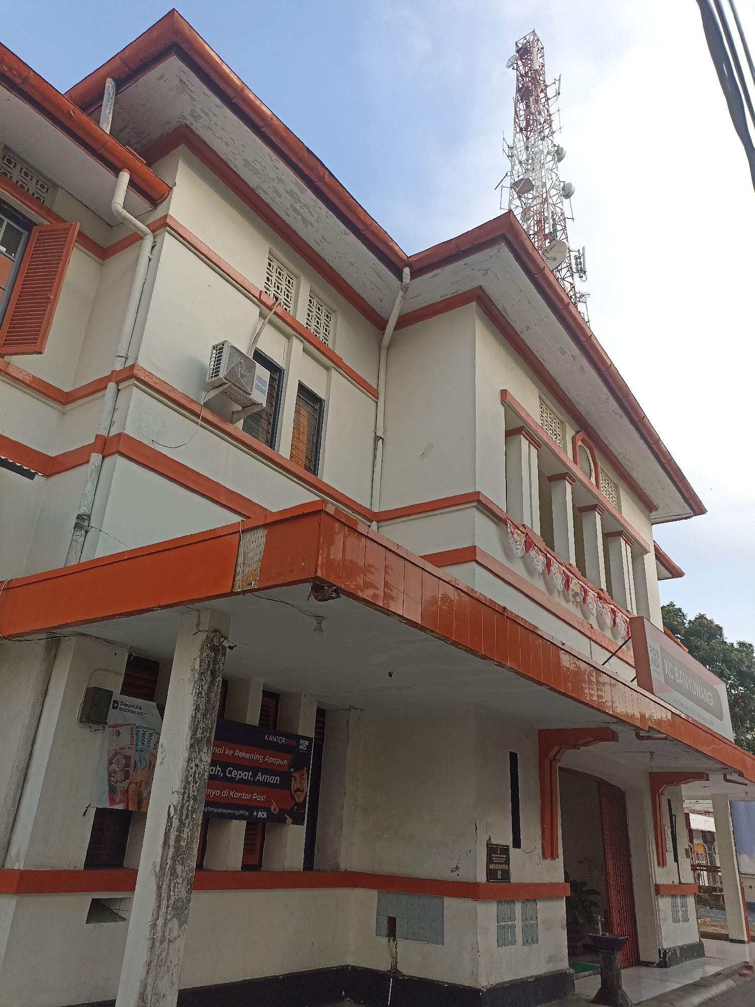 A low-angle, outdoor, medium shot captures the exterior of a white and orange building, possibly a post office, under a clear, pale blue sky. A prominent cell tower with red and white lattice work rises in the background. The building features orange trim along its roofline, overhangs, and window frames. Some windows are open with visible red shutters, while others have patterned grates. An air conditioning unit is mounted on the exterior wall. Several national flags, likely Indonesian, are strung along the building's facade. A sign on the building reads "KANTOR POS AD KC BANYUWANGI". Banners are visible on the building, one depicting Indonesian Rupiah currency. The ground in front of the building is paved. The building's facade shows signs of wear and tear, with peeling paint and some cracks in the plaster. The overall impression is of an older building in a somewhat dated state of repair, situated in an urban or semi-urban environment. The specific location is identified as Kelurahan Kepatihan, Indonesia.