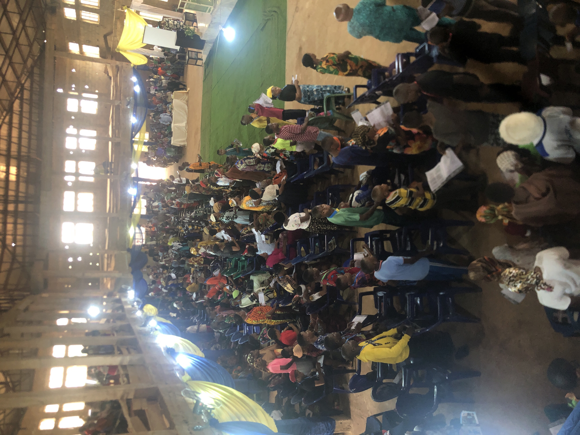 The image shows a large group of people gathered indoors in Obosi, Nigeria. They are seated in rows of blue plastic chairs, filling most of the space. The setting appears to be a large hall with a high ceiling, supported by beams and columns. Windows line the upper walls, allowing natural light to enter. Decorations of blue and yellow fabric are visible. At the far end, there is a raised platform covered in green material. The people appear to be observing or participating in an event.