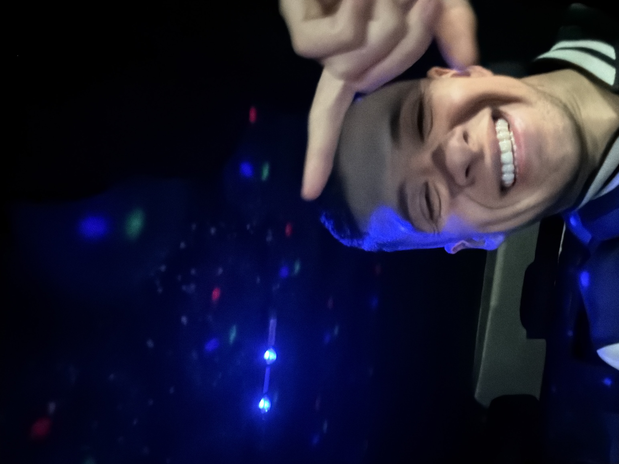 The image, rotated clockwise, features a person (appearing male) from the shoulders up, smiling broadly with white teeth, looking directly at the viewer. A blue light illuminates parts of their hair and forehead. They are wearing a dark top with a white striped collar or shoulder. The person's right hand is raised, with bent fingers, and the index finger is pointing downwards towards the left side of the frame.
The background is dark, resembling a nighttime or low-light indoor setting. Scattered across the dark background are numerous out-of-focus, bokeh-like points of light in blue, red, and green, indicative of disco or party lighting. More distinct blue light sources are visible in the lower left portion of the image. A dark, rectangular object with a light edge is partially visible along the right side. The overall mood is joyful and energetic, typical of a social gathering or event. No text is visible.