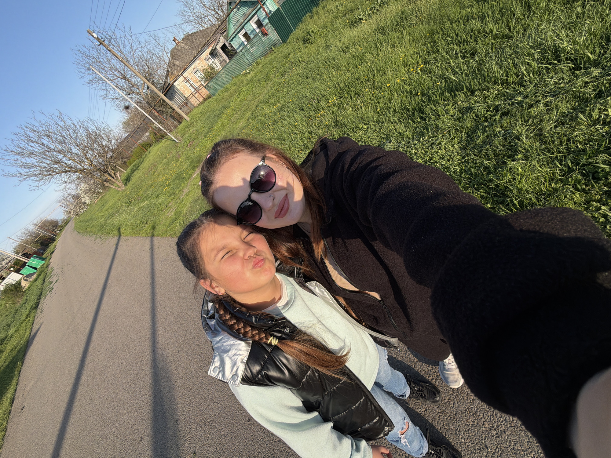 A tilted outdoor selfie features two females, an adult/teenager and a younger girl, posing together on a sunny day in what appears to be Novopokrovskaya, Russia.
The adult/teenager, positioned closer to the camera and holding it, has brown hair and wears dark, round sunglasses that reflect the bright sky. She is dressed in a black hooded jacket and has light-colored lipstick, smiling gently towards the camera. Her face is partially shadowed by the sunglasses.
Beside her, the younger girl makes a playful pouting face with her eyes closed. Her brown hair is styled in long braids. She is dressed in a light blue long-sleeved top, a shiny silver and black puffer vest, distressed blue jeans, and dark shoes.
The setting is a rural or semi-rural landscape under a clear blue sky. To the left, a paved road stretches, showing long shadows from what appear to be utility poles, suggesting either early morning or late afternoon. To the right, a grassy embankment slopes upwards, featuring lush green grass and scattered yellow dandelions. In the mid-ground, an old, weathered house with a distinct roofline and brick/stone facade is visible, partially obscured by a green metal fence and bare trees, one of which displays white blossoms, hinting at springtime. Power lines crisscross the sky near a wooden utility pole.
The overall mood is cheerful and relaxed, capturing a moment between the two individuals enjoying the outdoors. No visible text is present in the image.